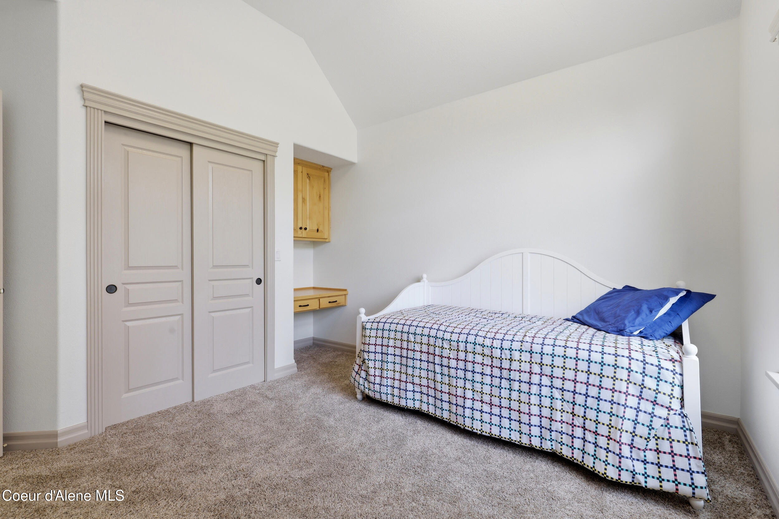 Bedroom with a white single bed, colorful checkered bedding, and built-in wooden desk beside a double-door closet.
