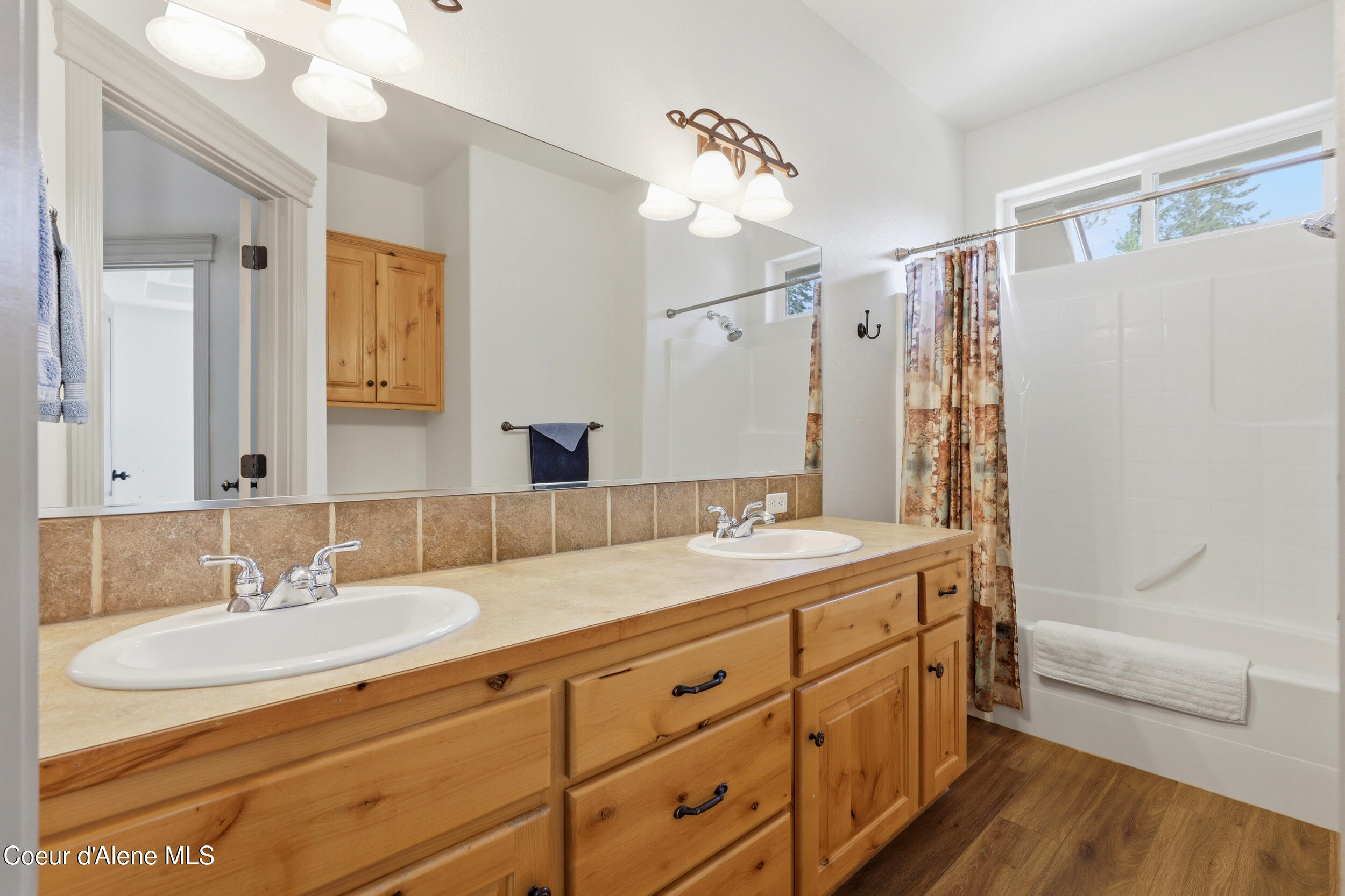 Bathroom with a long honey-colored double vanity, large mirror, and tub-shower with patterned curtain.
