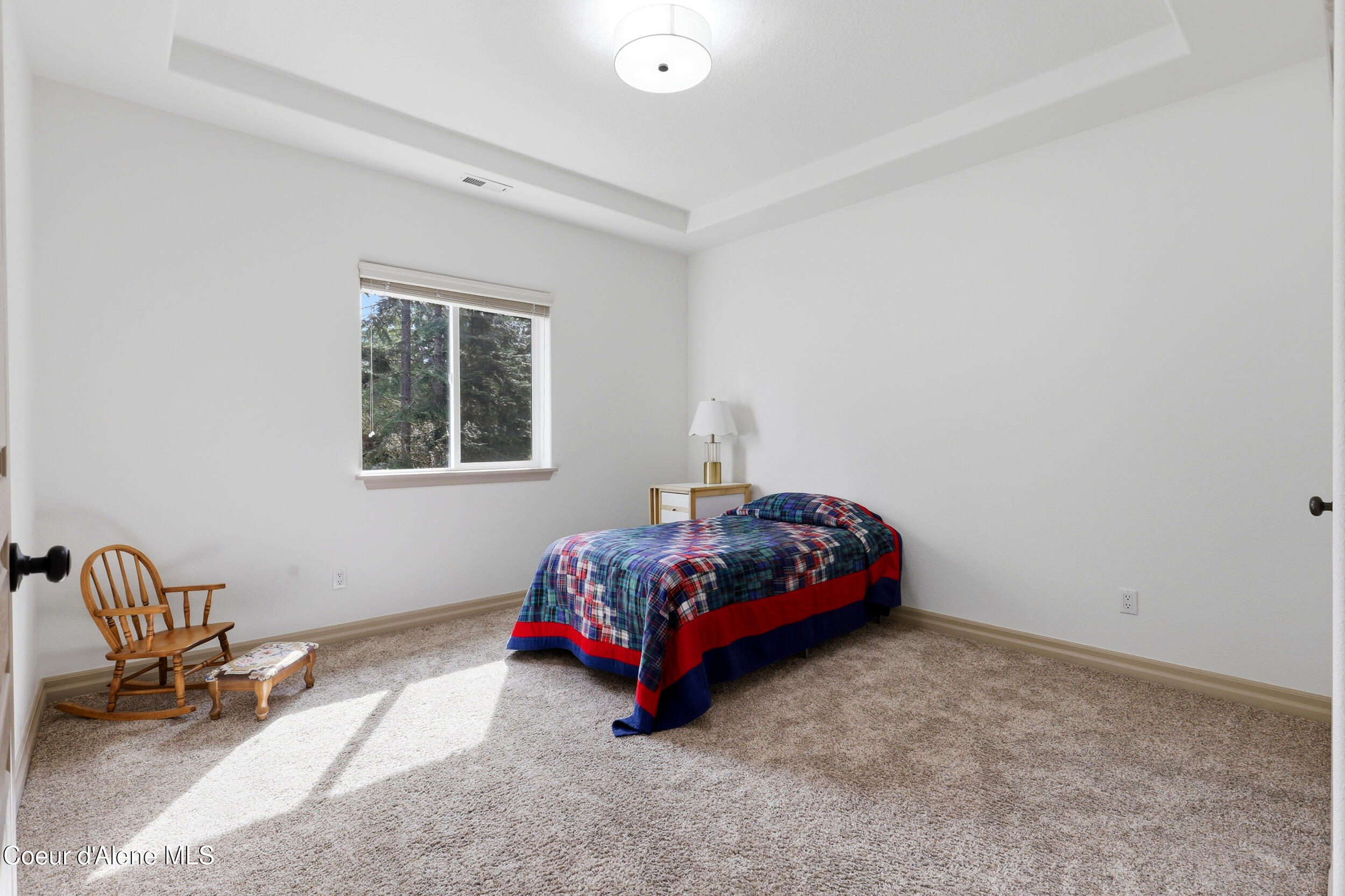 Furnished bedroom with a plaid-covered bed, rocking chair, and sunlit window overlooking trees.