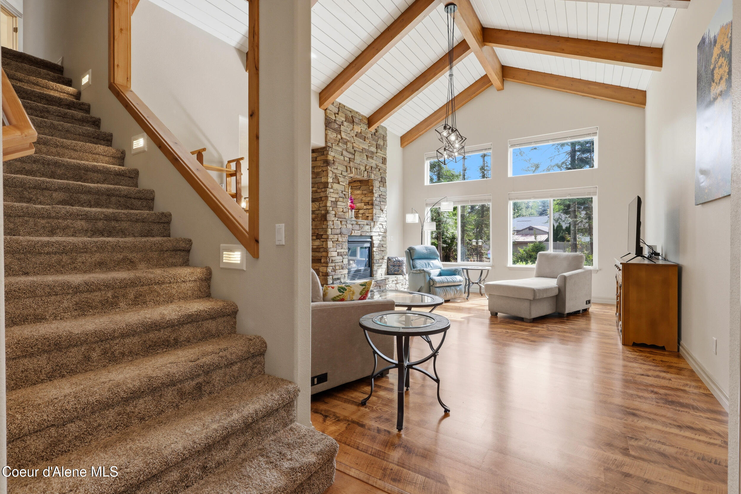 Vaulted living room with exposed beams, stone fireplace, large windows, and carpeted staircase.