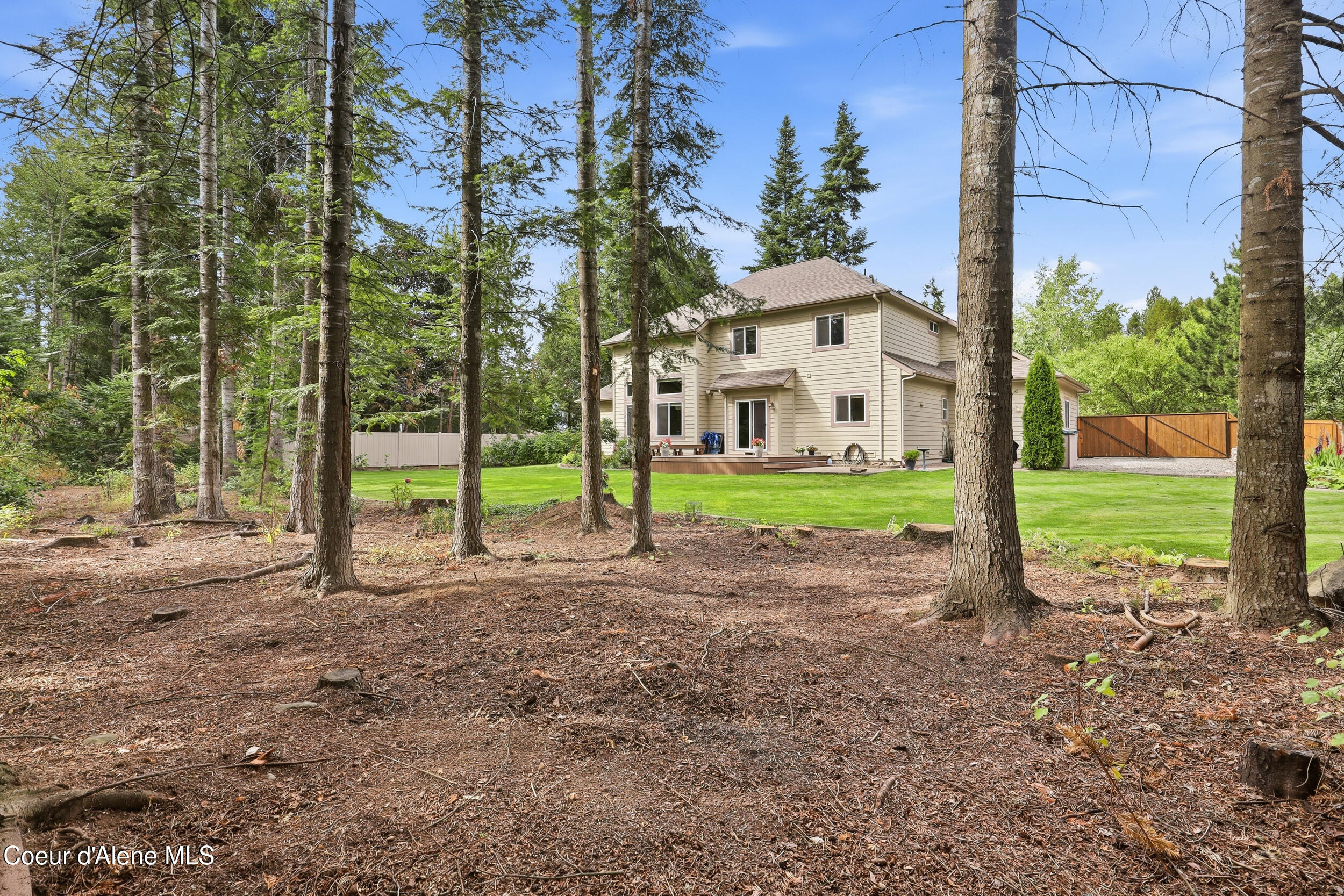 Two-story beige home beyond a lawn, framed by tall evergreens and a wooden driveway gate.