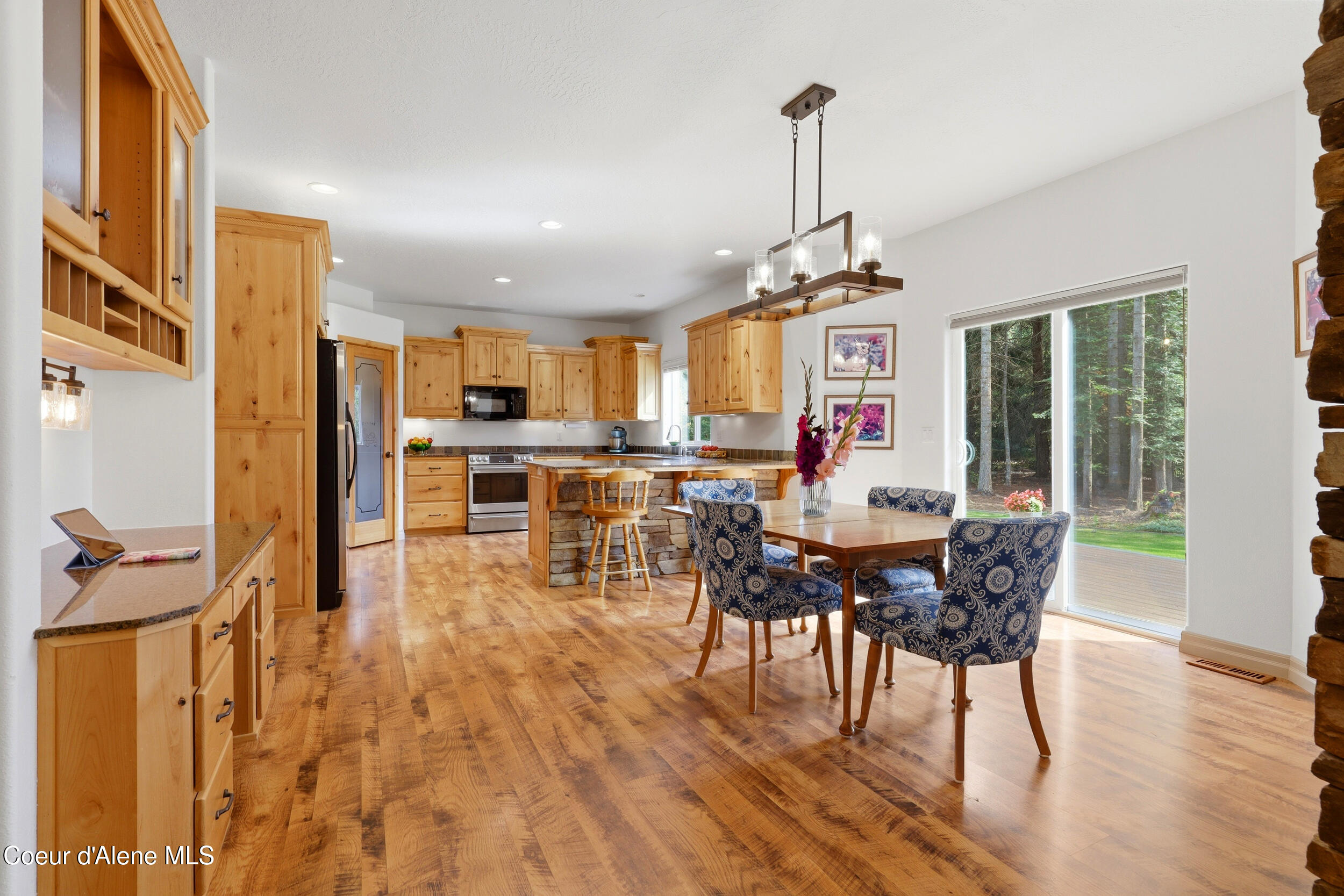 Open kitchen and dining area with honey-toned cabinetry, a stone island, and sliding doors to a wooded yard.