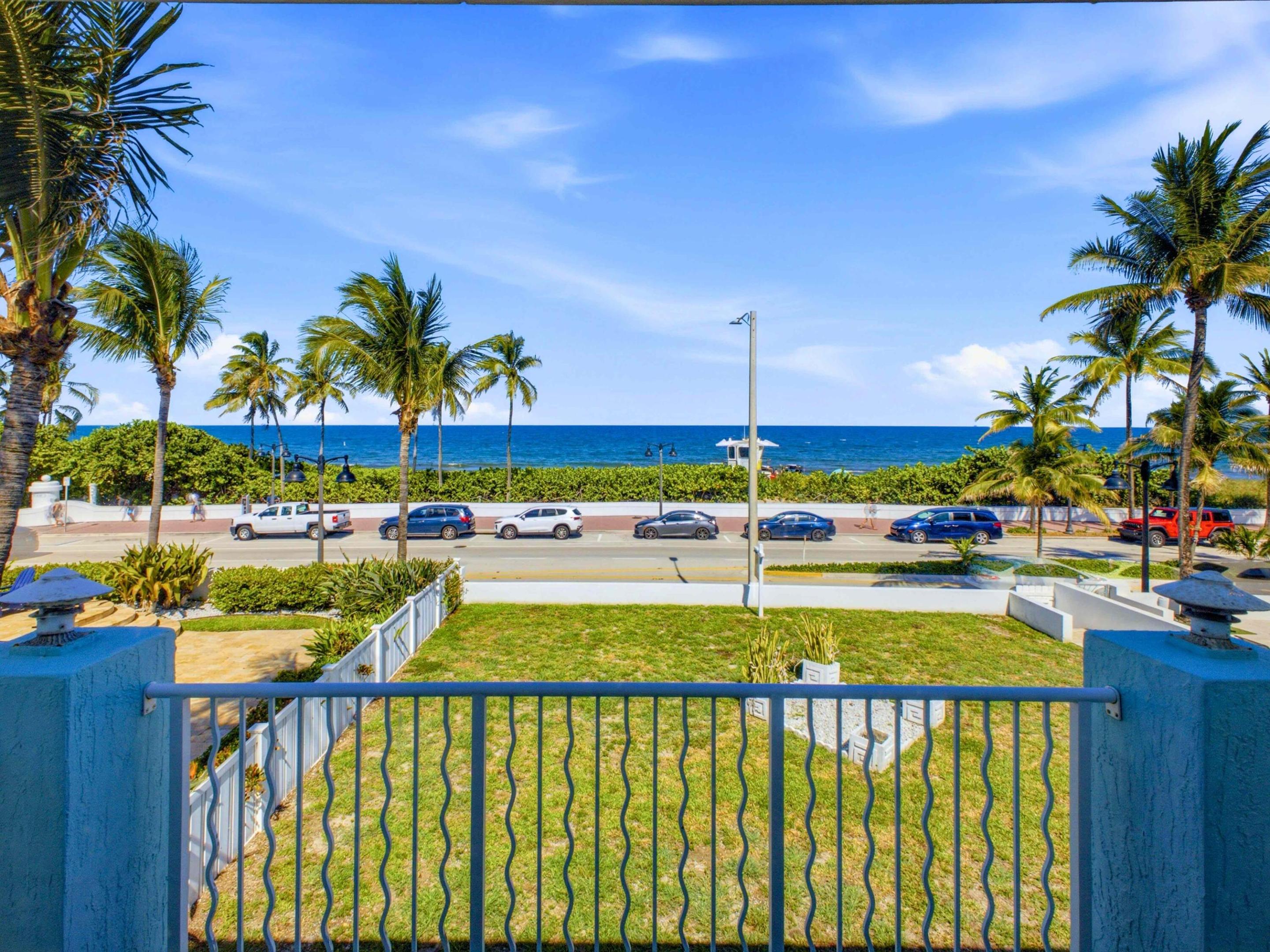 Ocean view beyond a palm-lined road, seen from a fenced lawn with a blue railing.