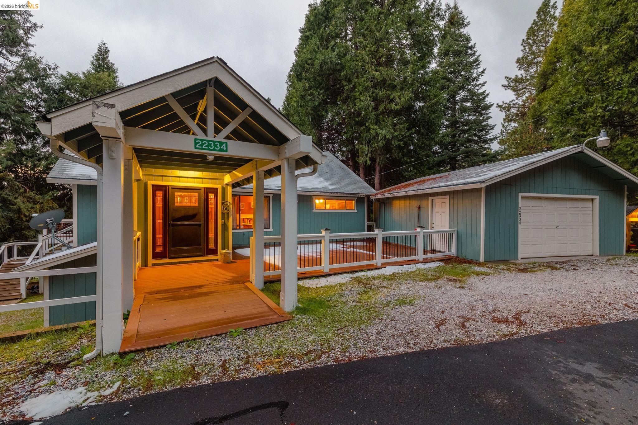 Teal-sided home with a broad covered porch, warmly lit entrance, and garage among tall evergreens.