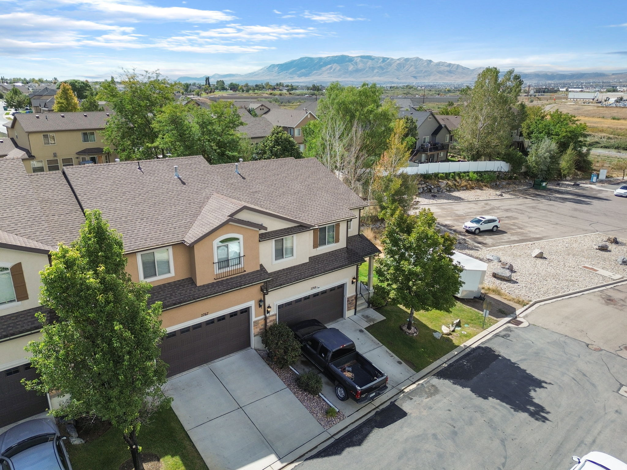 Aerial view of a tan two-story home with two garages, mature trees, and mountains beyond the neighborhood.