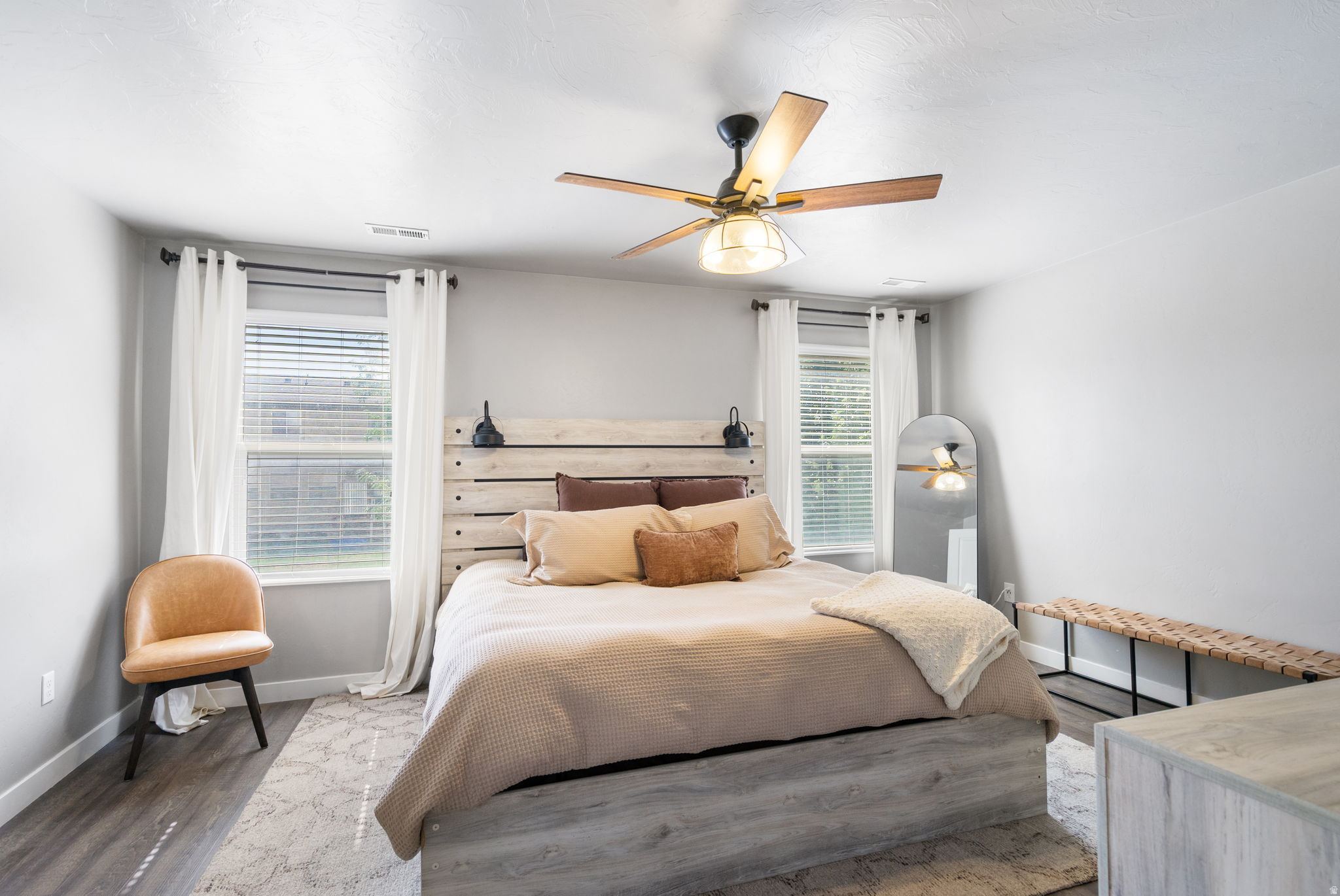 Staged bedroom with a plank headboard, two curtained windows, and a wood-blade ceiling fan.