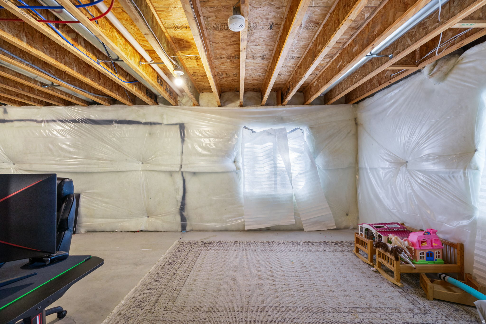 Unfinished basement with exposed ceiling joists, insulated walls, a rug, and children's toys.