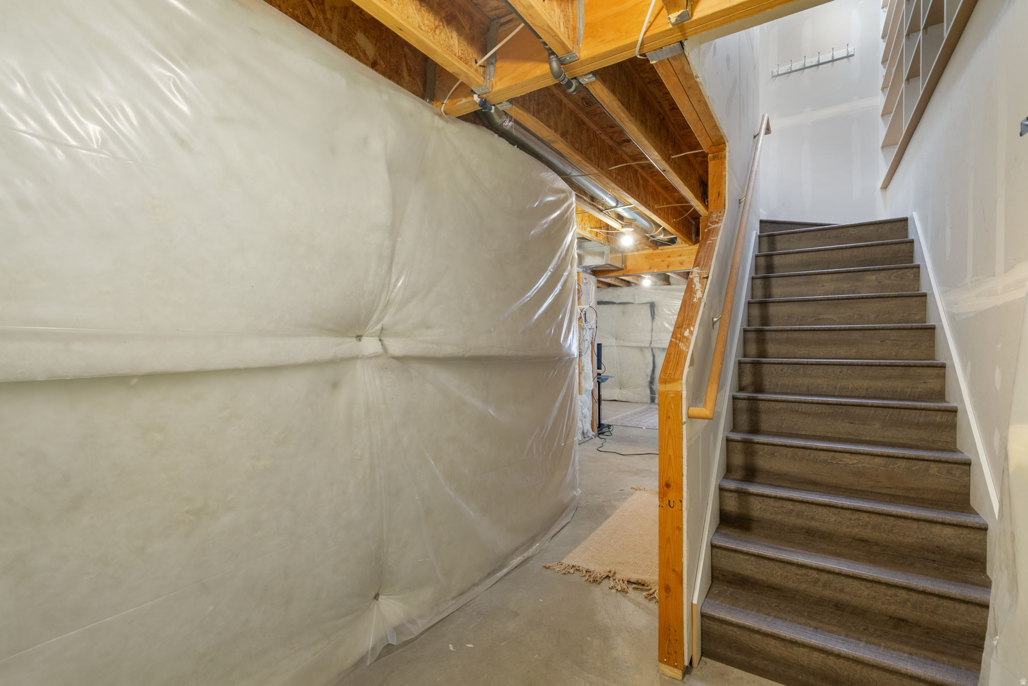Basement stairwell with carpeted steps, a wood handrail, exposed ceiling joists, and plastic-covered insulation.