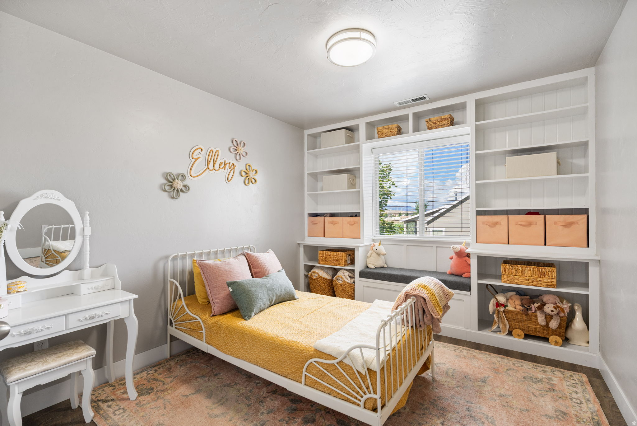 Child's bedroom with a white metal bed, mustard bedding, built-in shelves, and a window seat.