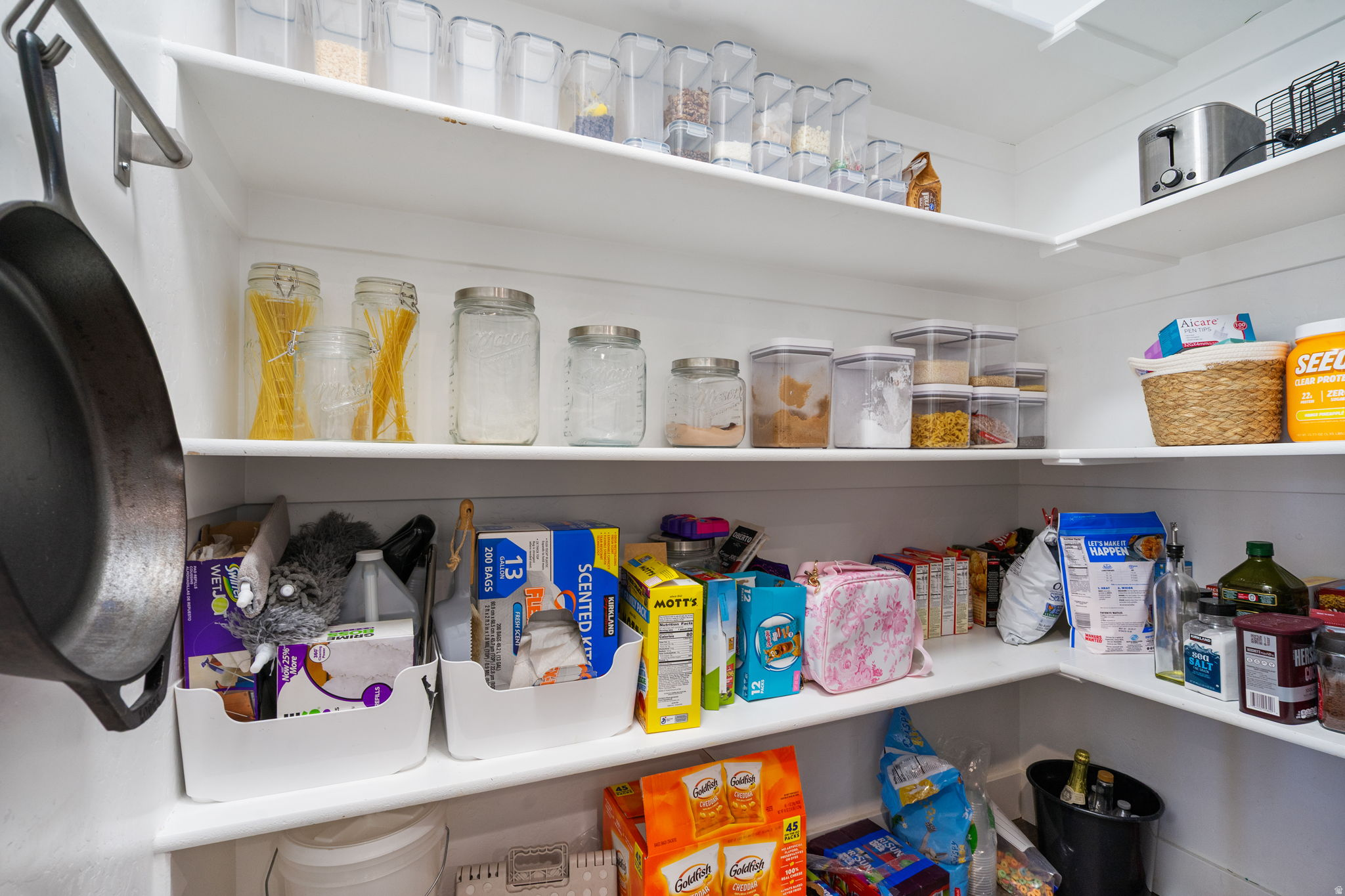 Well-stocked pantry with wraparound white shelves, clear storage jars, baskets, and packaged food.