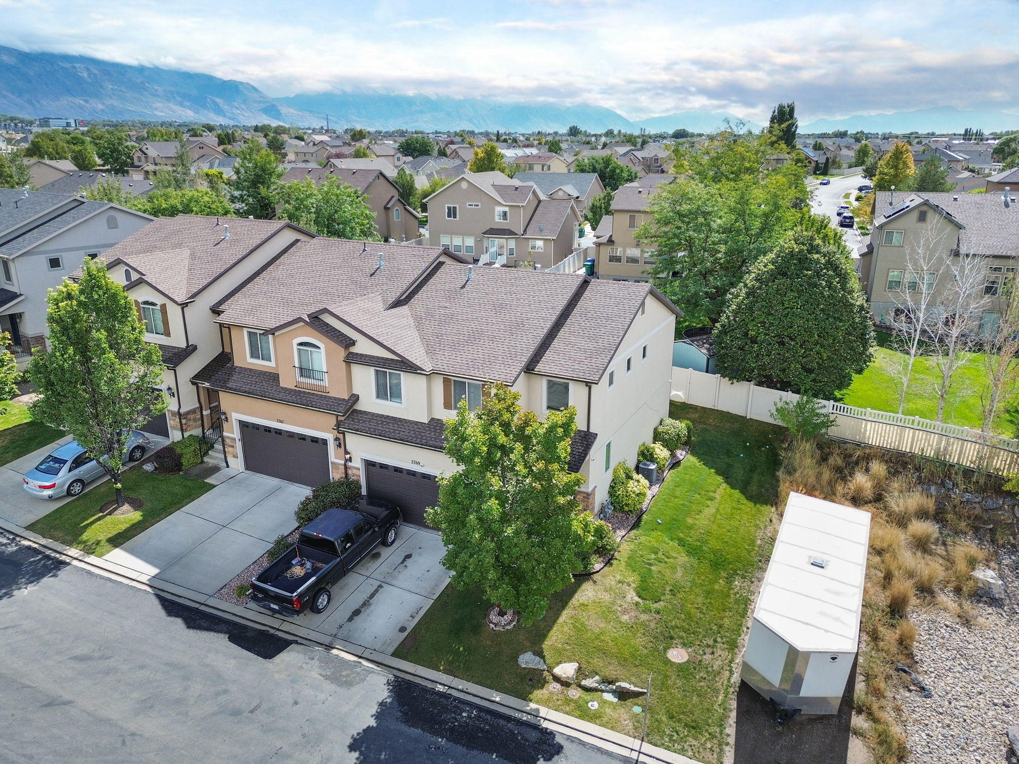 Aerial view of a beige two-story home with a two-car garage, lawn, fenced yard, and mountain backdrop.