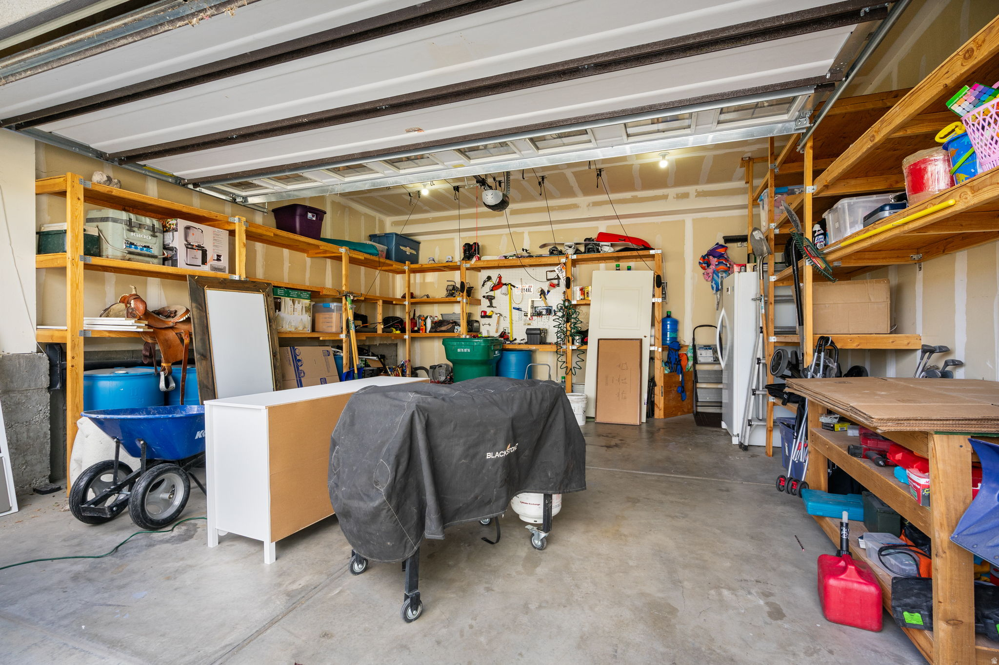 Garage with open overhead door, extensive wooden shelving, tools, and a covered Blackstone grill.