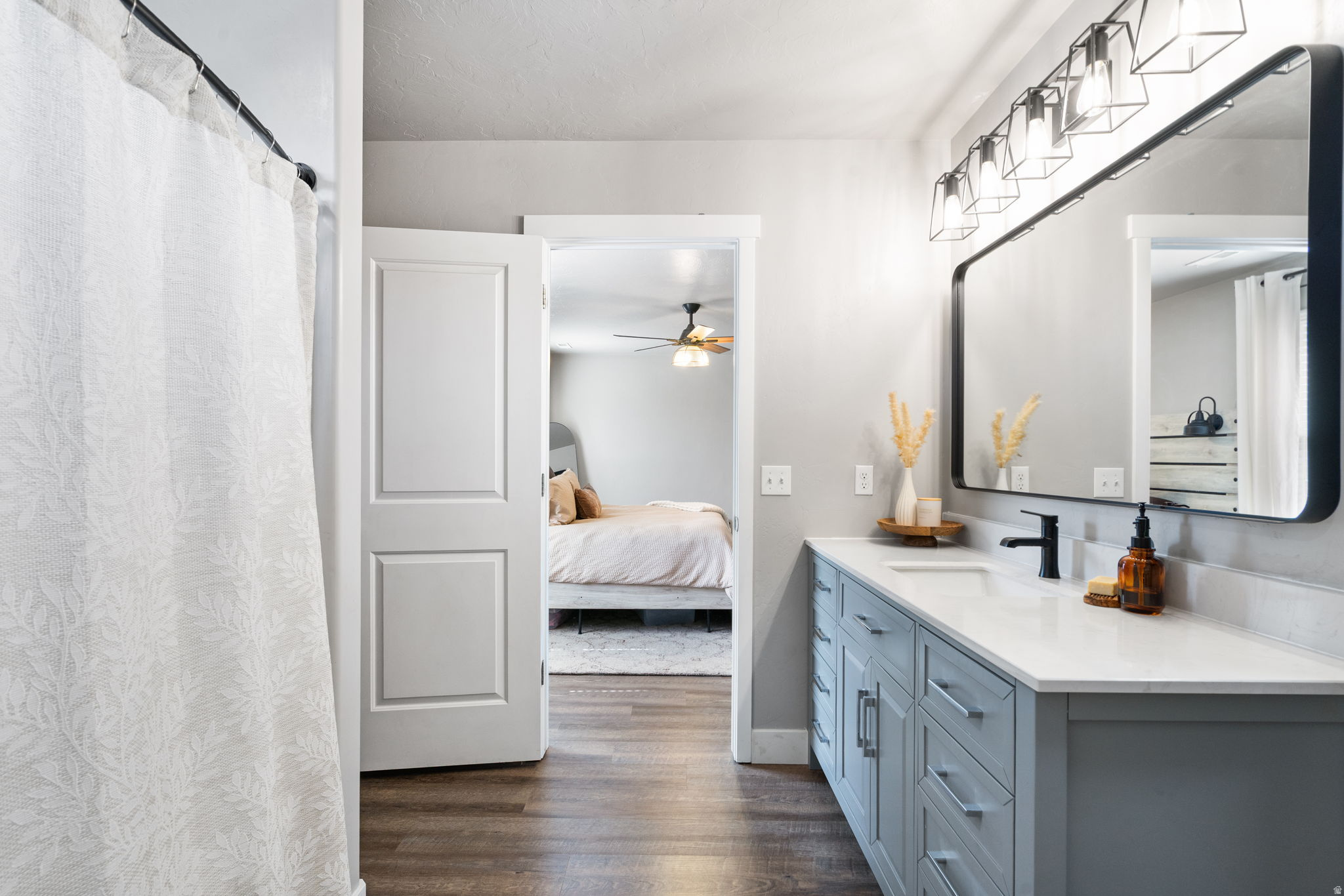Bathroom with a blue-gray vanity and black-framed mirror beside an open doorway to a bedroom.