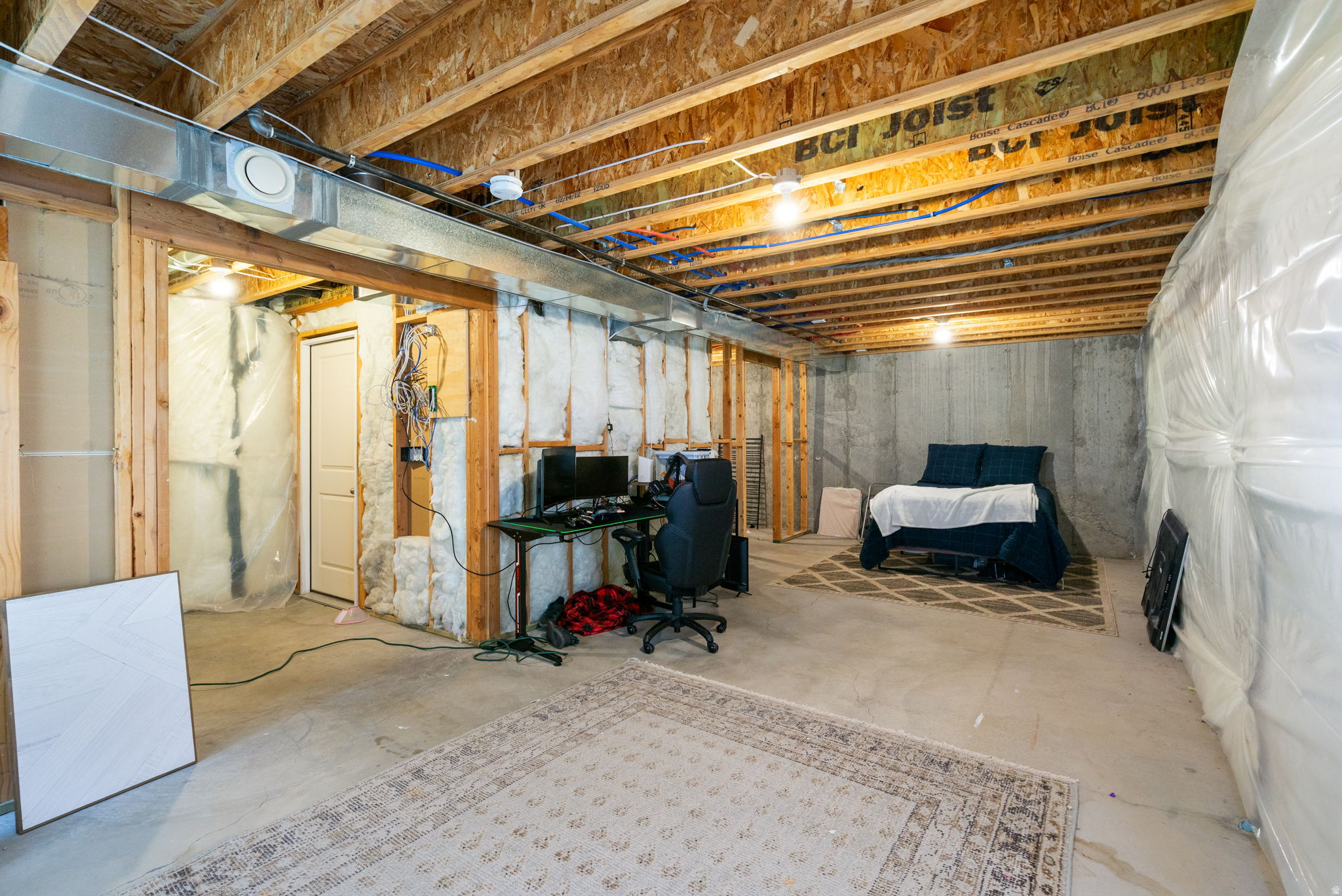 Unfinished basement with exposed framing, a computer workstation, and a bed on a rug.