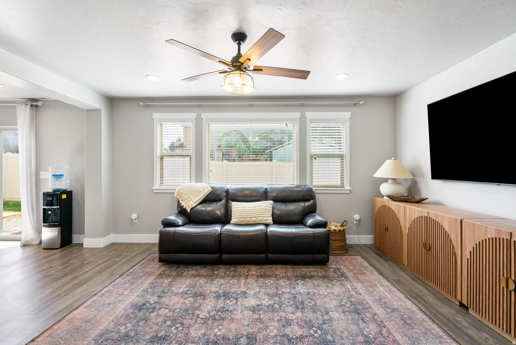 Furnished living room with a black leather sofa, three windows, and a patterned rug.