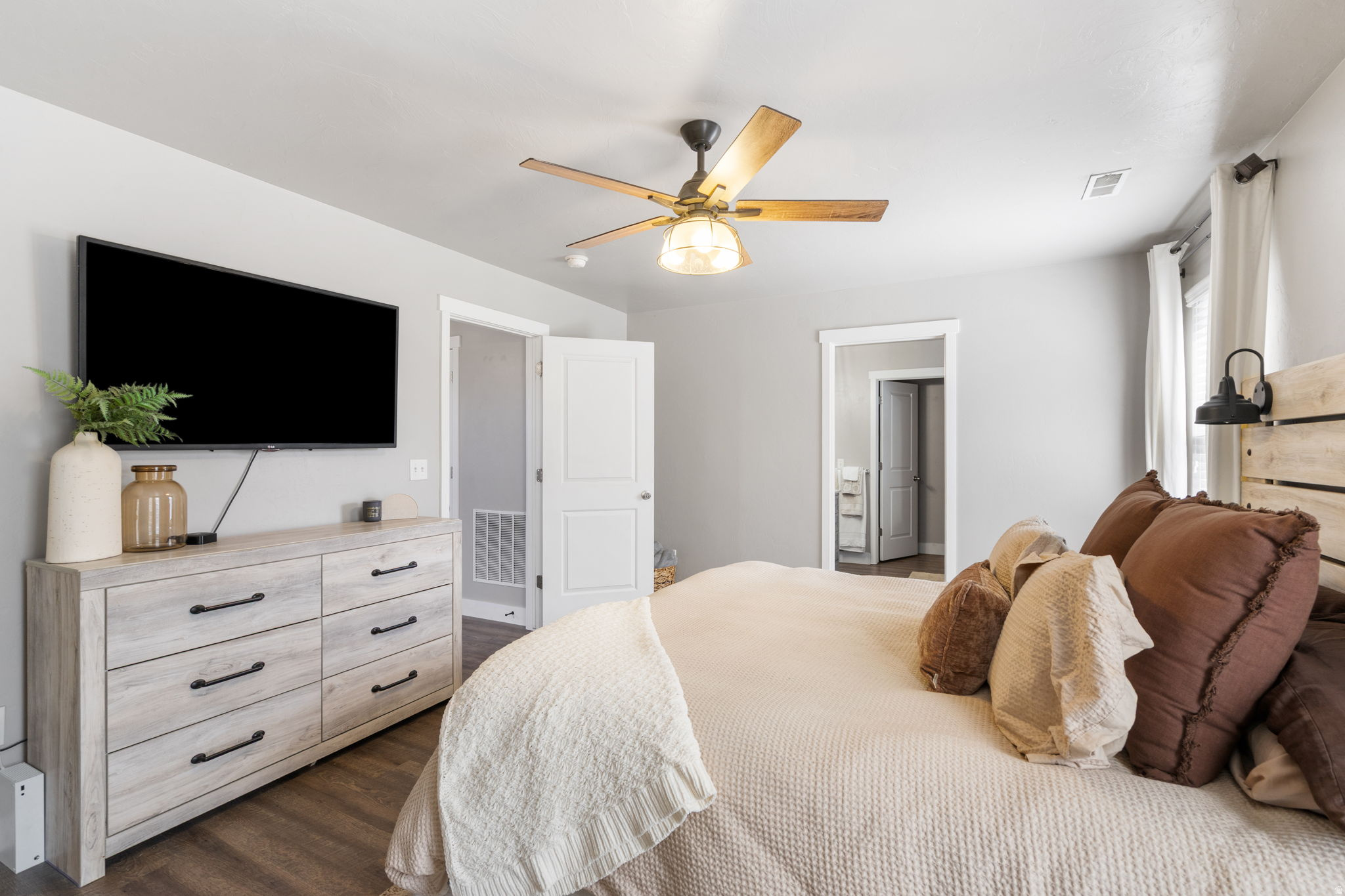 Staged bedroom with cream bedding, brown pillows, a wall-mounted TV, and a ceiling fan.