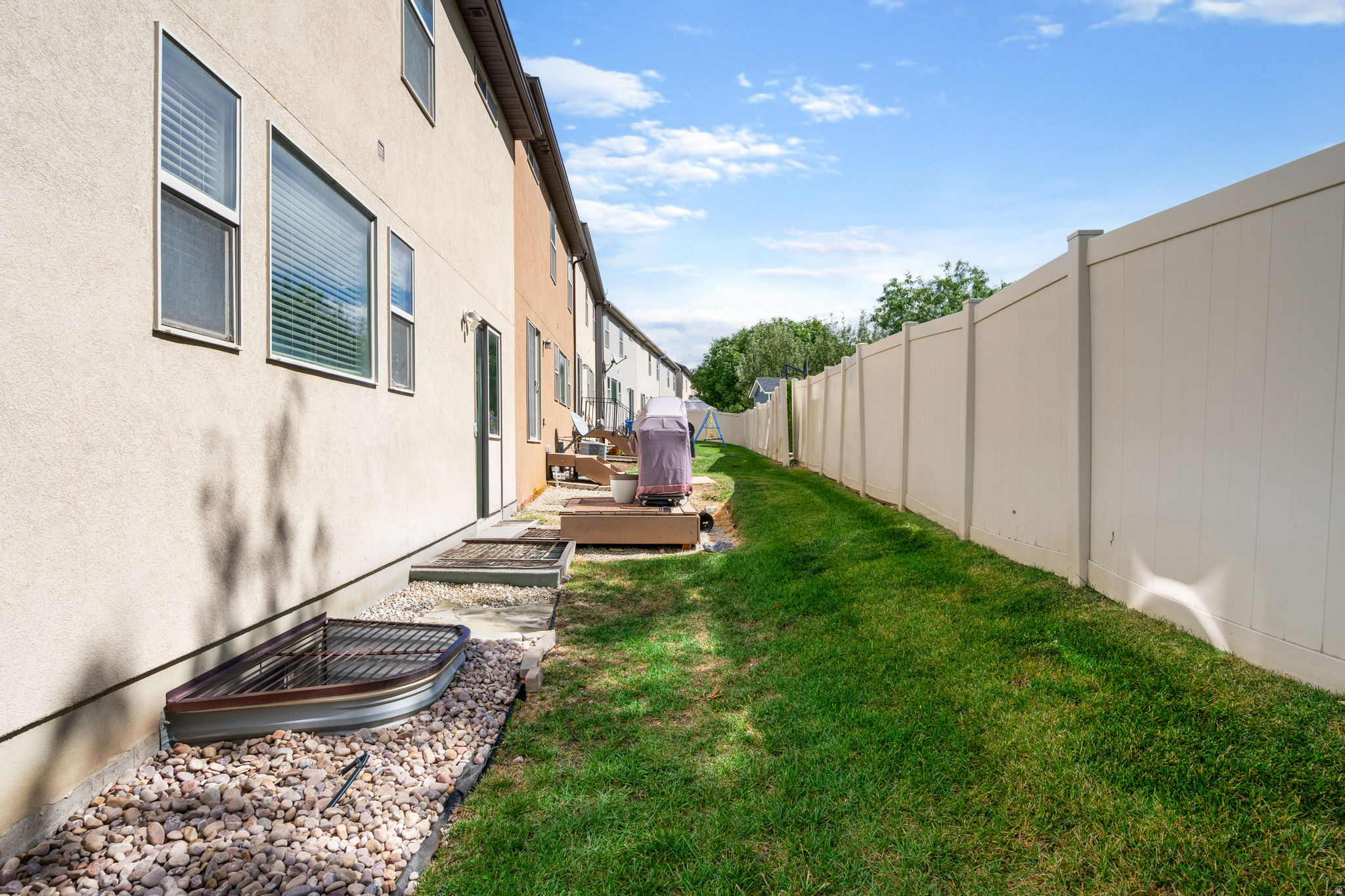 Side yard with a long lawn, white privacy fence, and rock landscaping beside the stucco home.