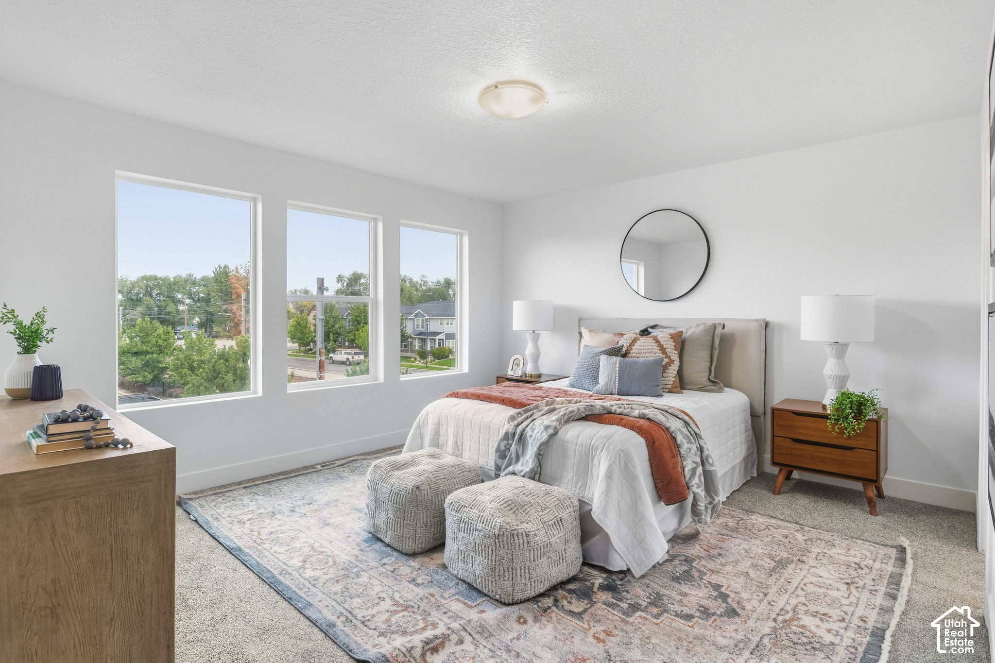Staged bedroom with three large windows, a white bed, knitted ottomans, and a patterned rug.
