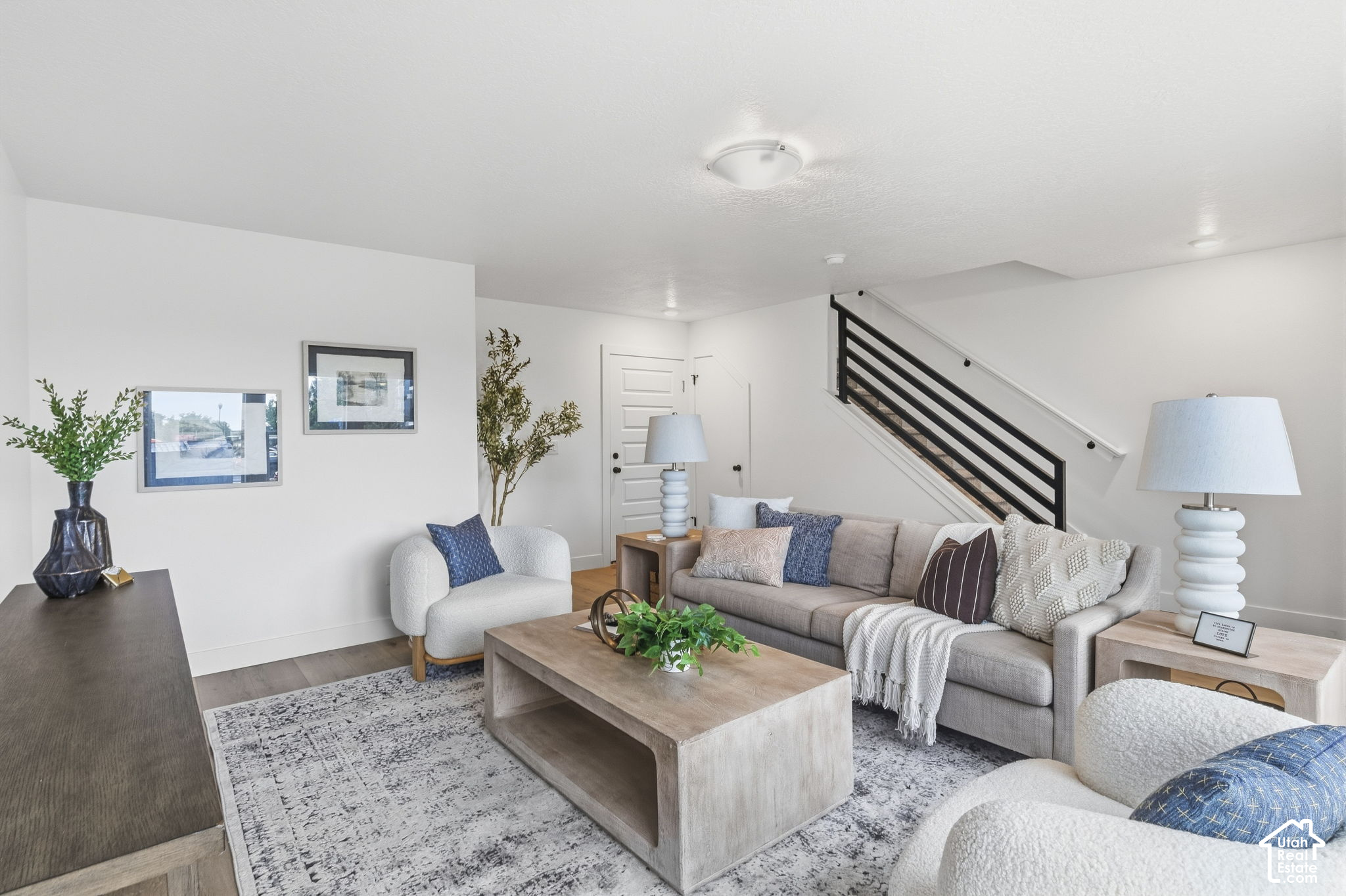 Staged living room with gray sofa, white armchairs, light wood coffee table, and black-railed staircase.