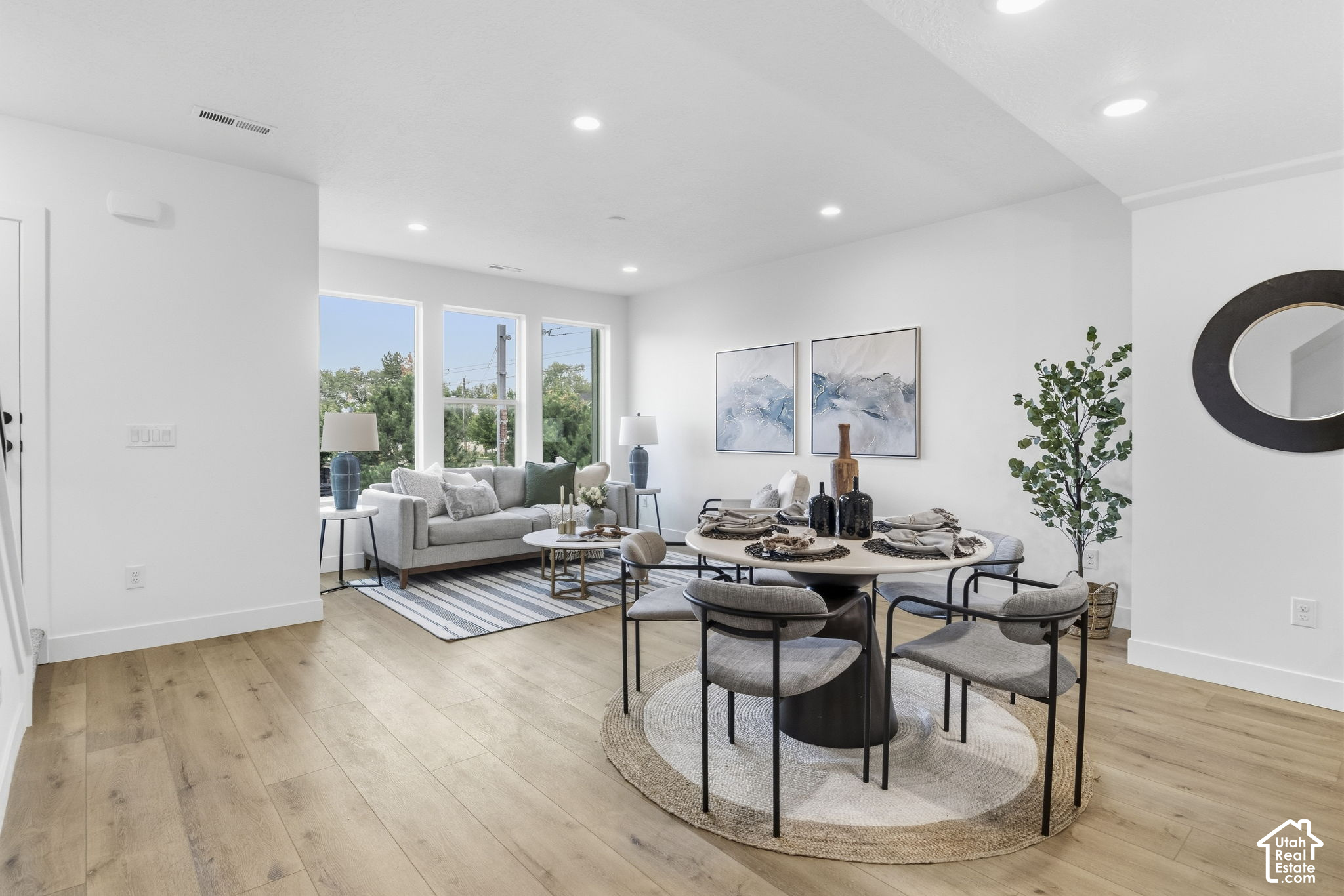 Staged dining and living room with a round table, gray sofa, large windows, and light wood floors.