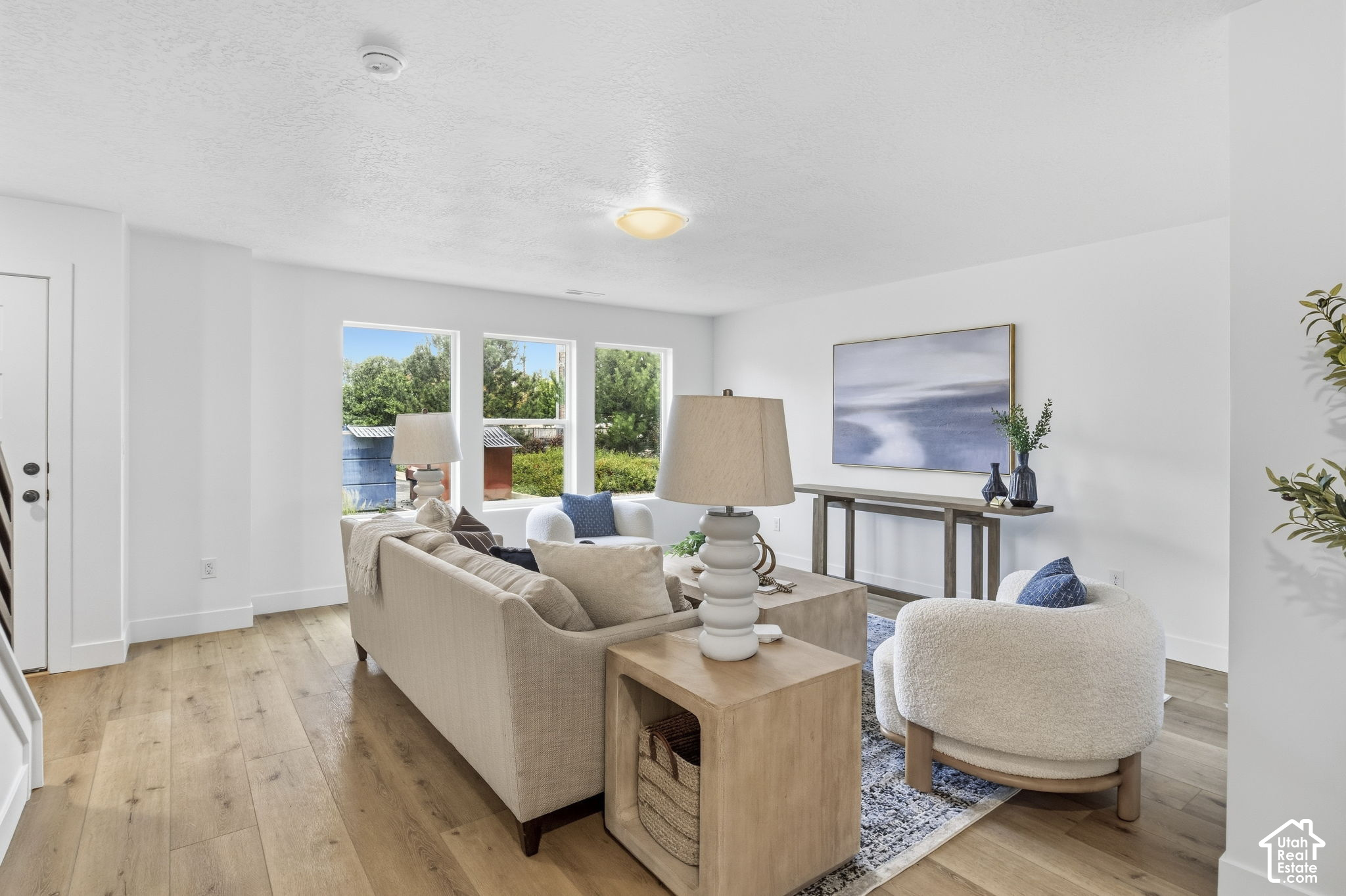 Staged living room with neutral seating, light wood floors, and three windows overlooking greenery.