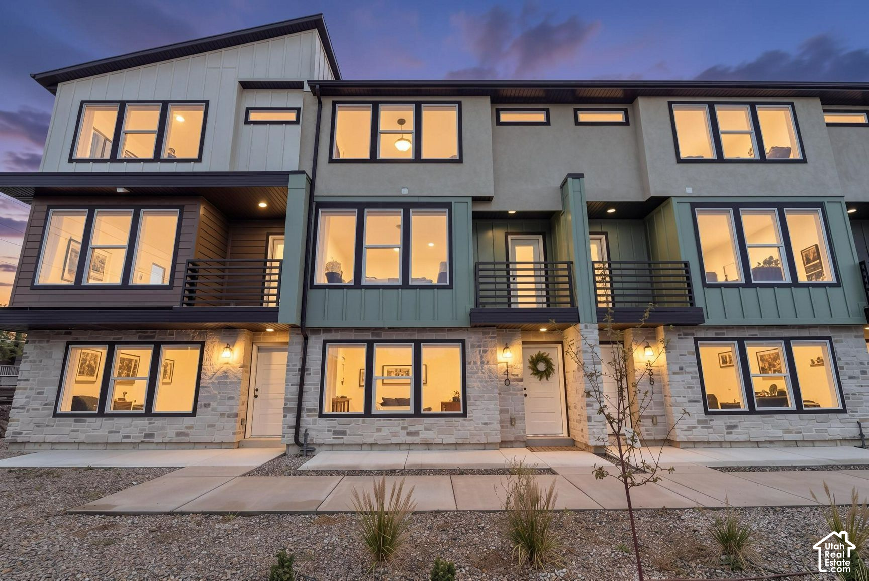 Modern multi-unit building with stone ground floor, balconies, and warmly lit windows at dusk.