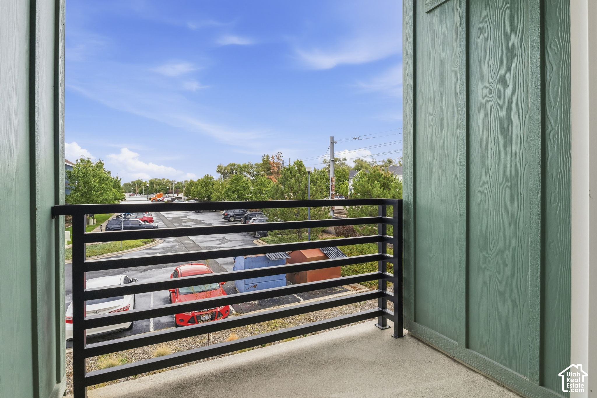 Covered balcony with green siding and black railing overlooking a parking lot and trees.