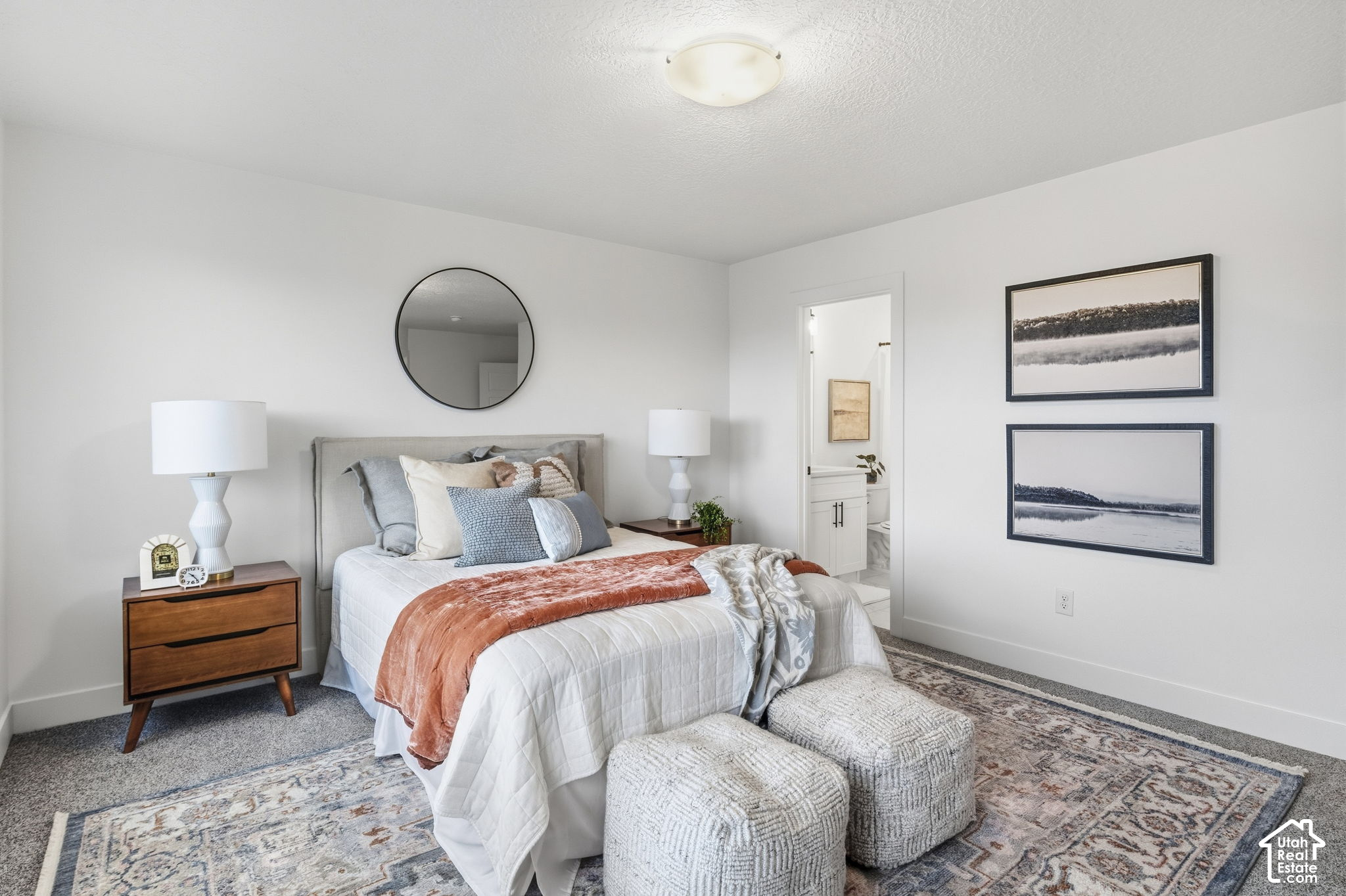 Staged bedroom with a gray upholstered bed, rust-colored throw, textured ottomans, and ensuite bathroom doorway.