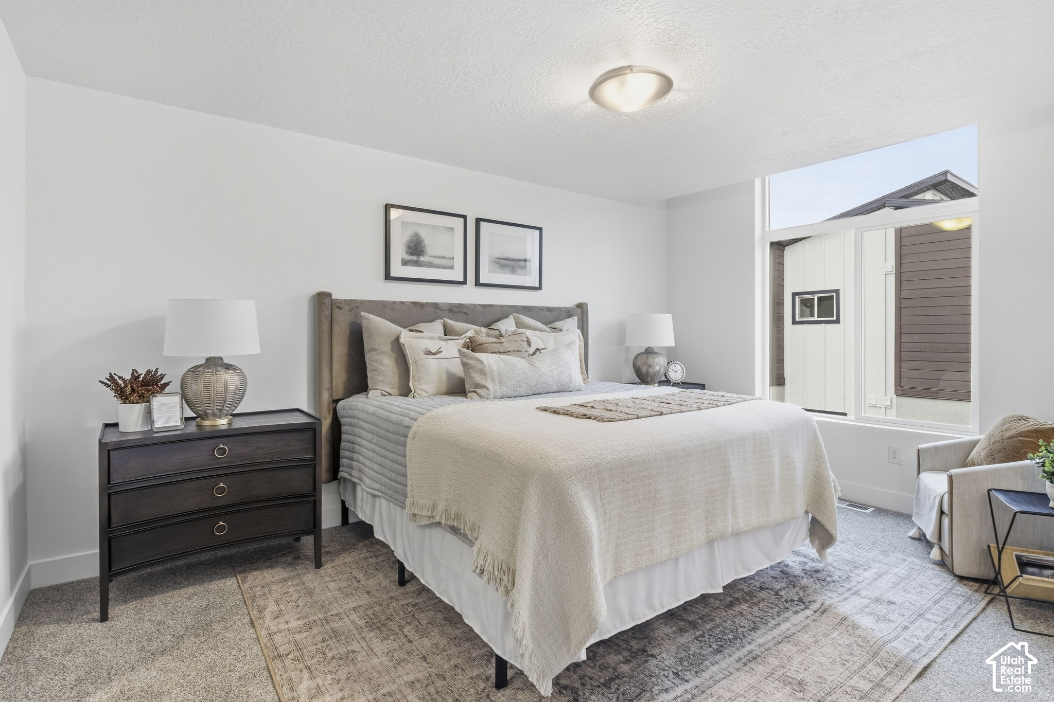 Staged bedroom with a large neutral bed, dark dresser, and wide window.