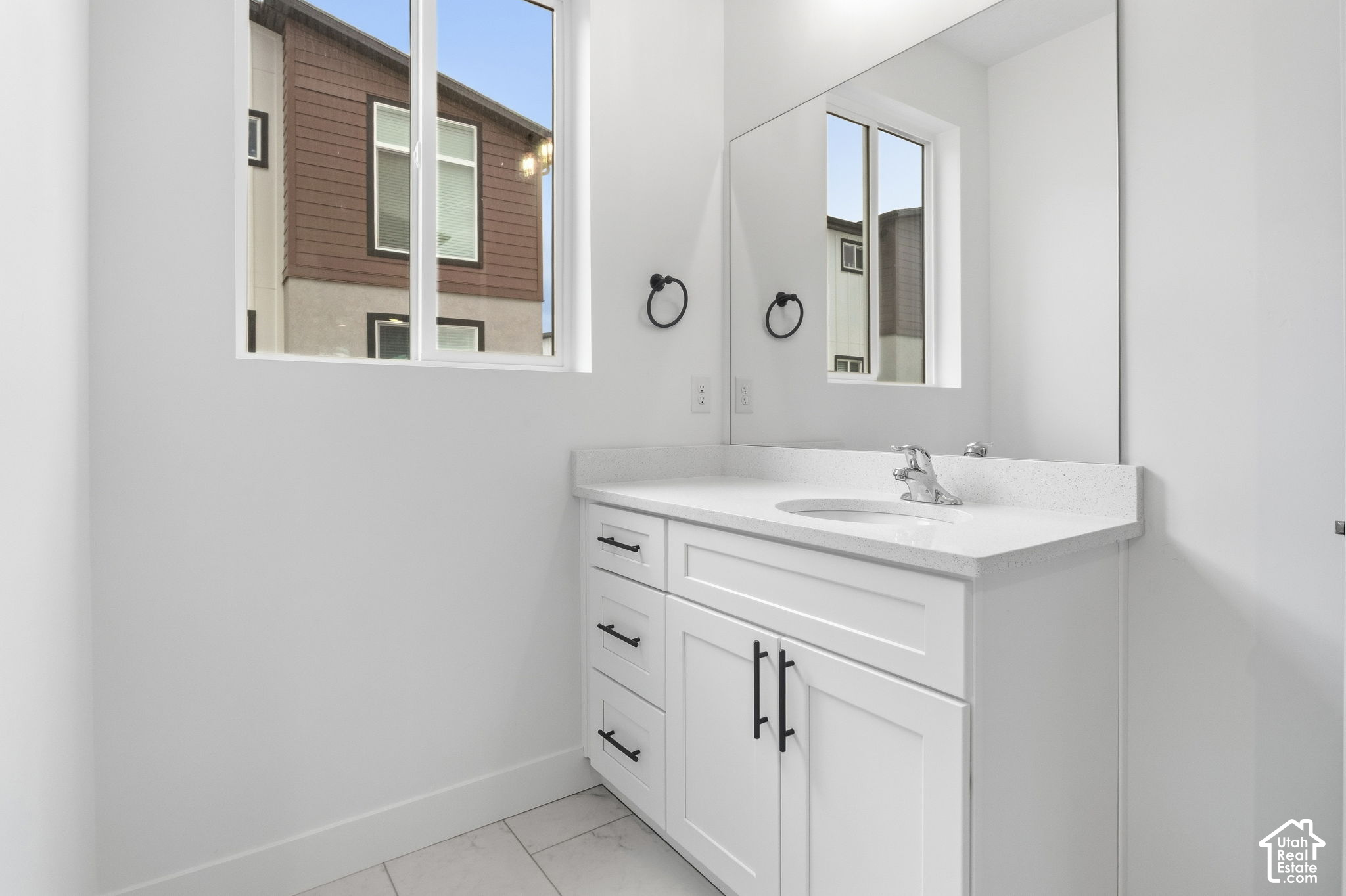 Bathroom vanity with white cabinetry, speckled counter, large mirror, and a window overlooking a neighboring building.