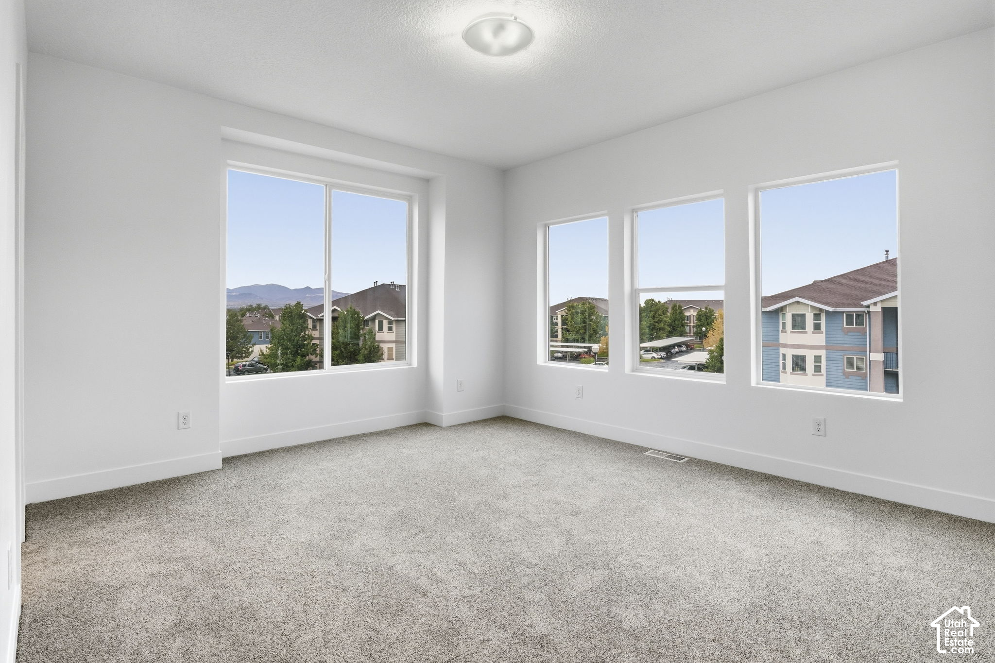 Empty carpeted room with large windows overlooking neighboring homes and distant mountains.