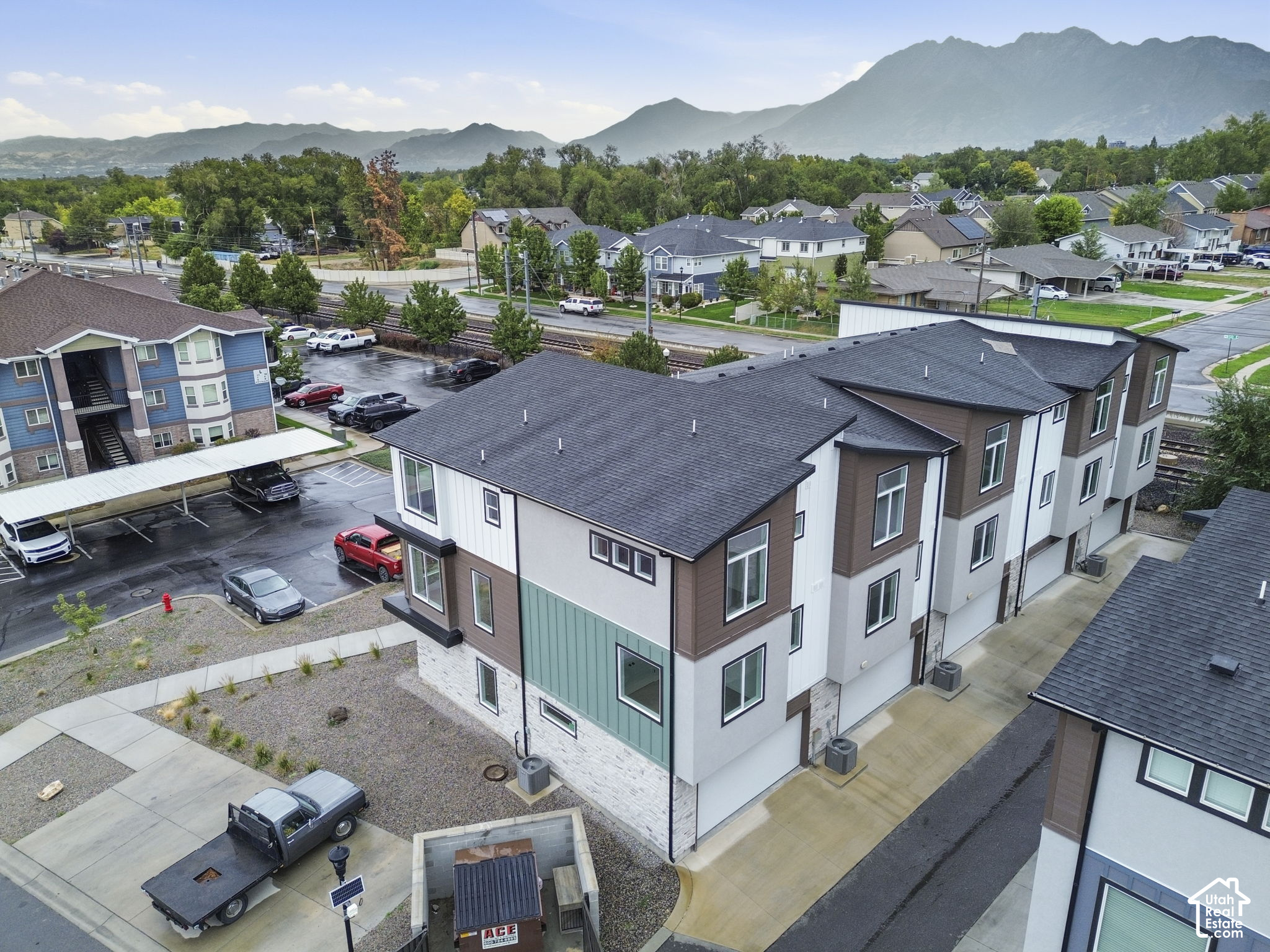 Modern multi-unit building with teal siding, parking, nearby homes, and mountains in the background.