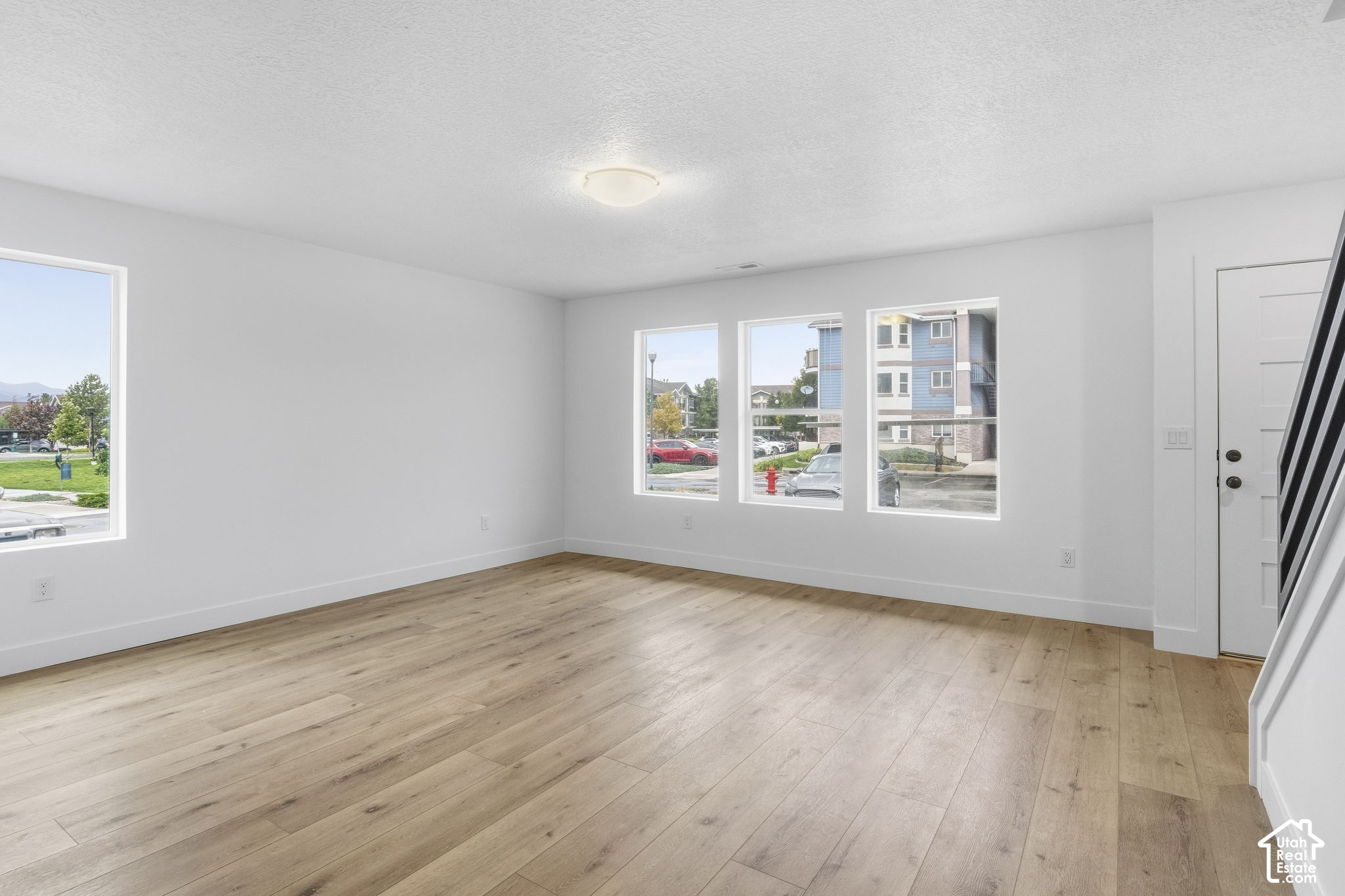 Vacant room with light wood floors, large windows, and a staircase.