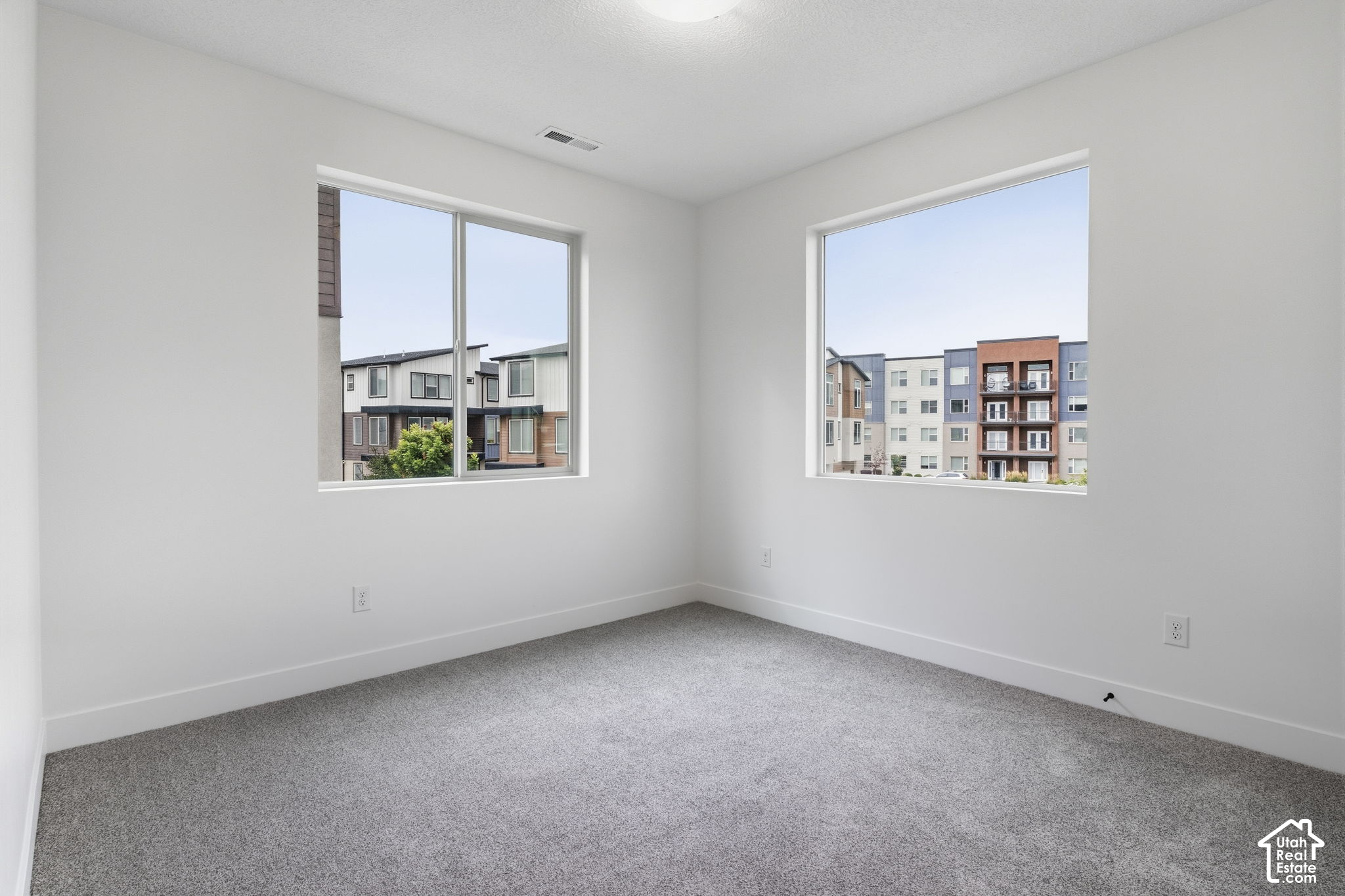 Empty carpeted room with two windows overlooking neighboring apartment buildings.