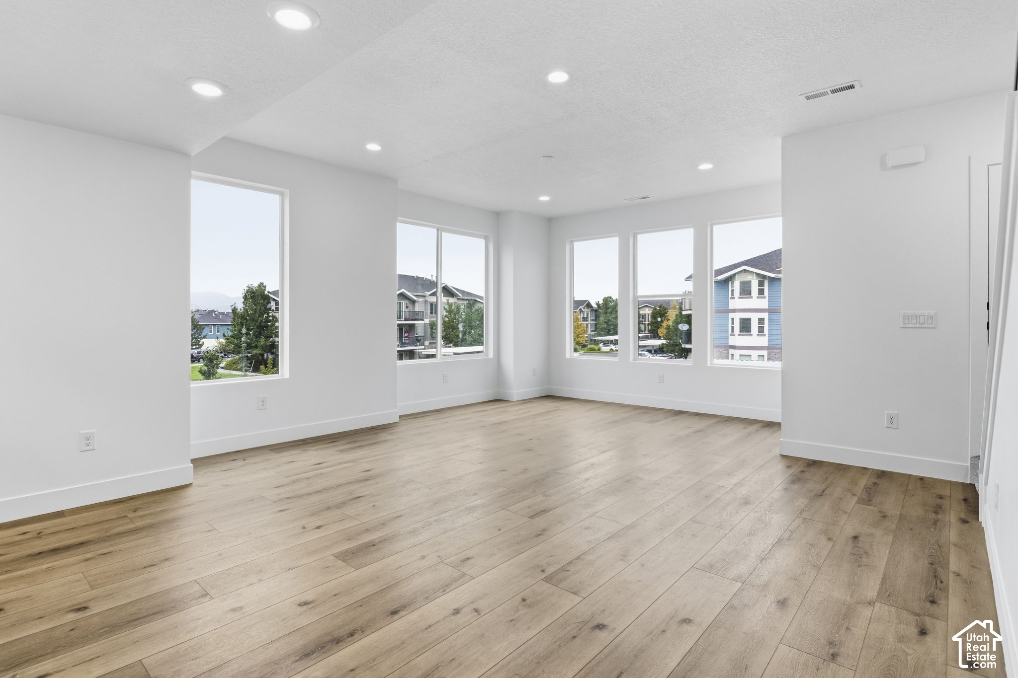 Empty living area with light wood floors and large windows overlooking nearby homes.