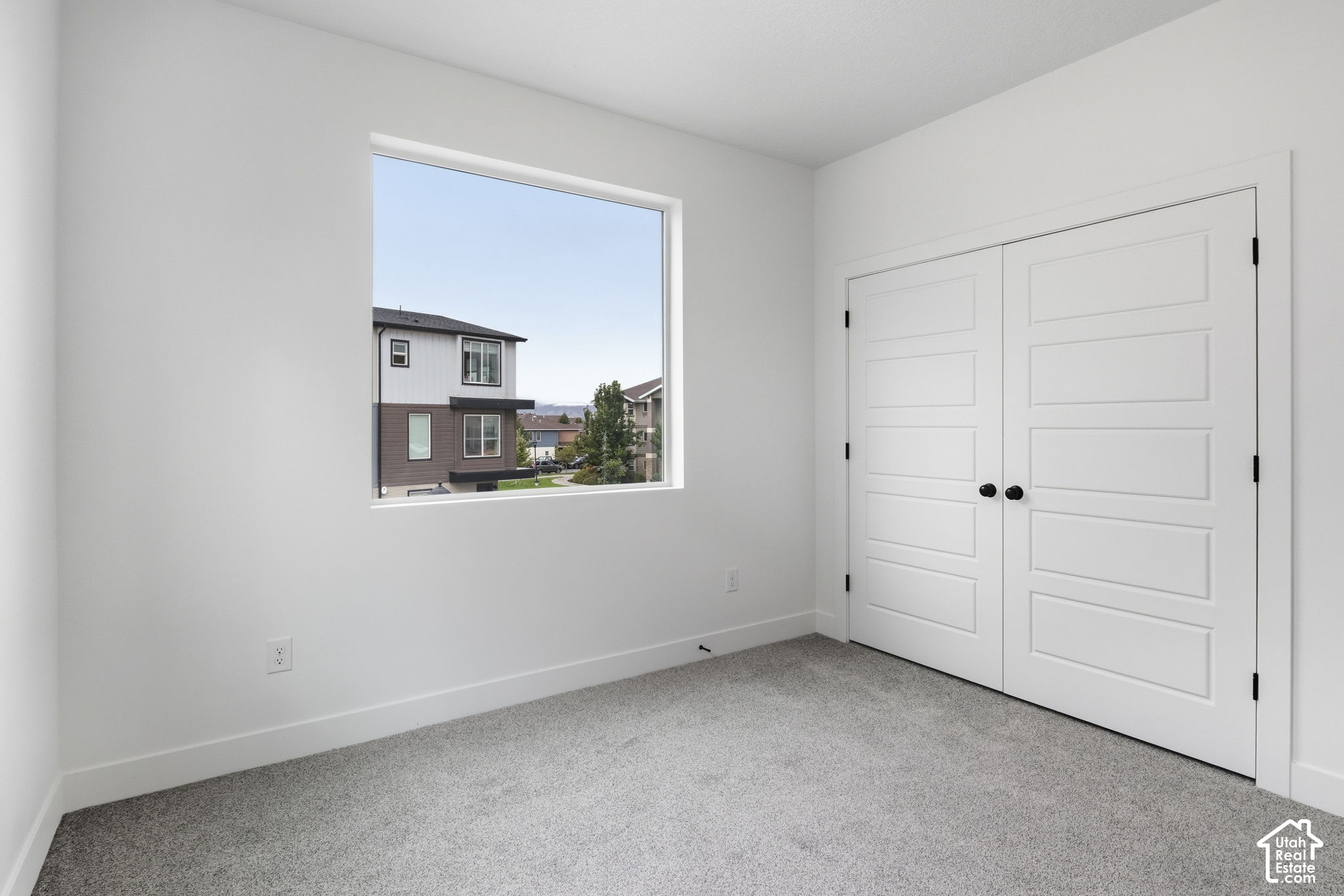 Empty bedroom with gray carpet, white double closet doors, and a window overlooking neighboring homes.