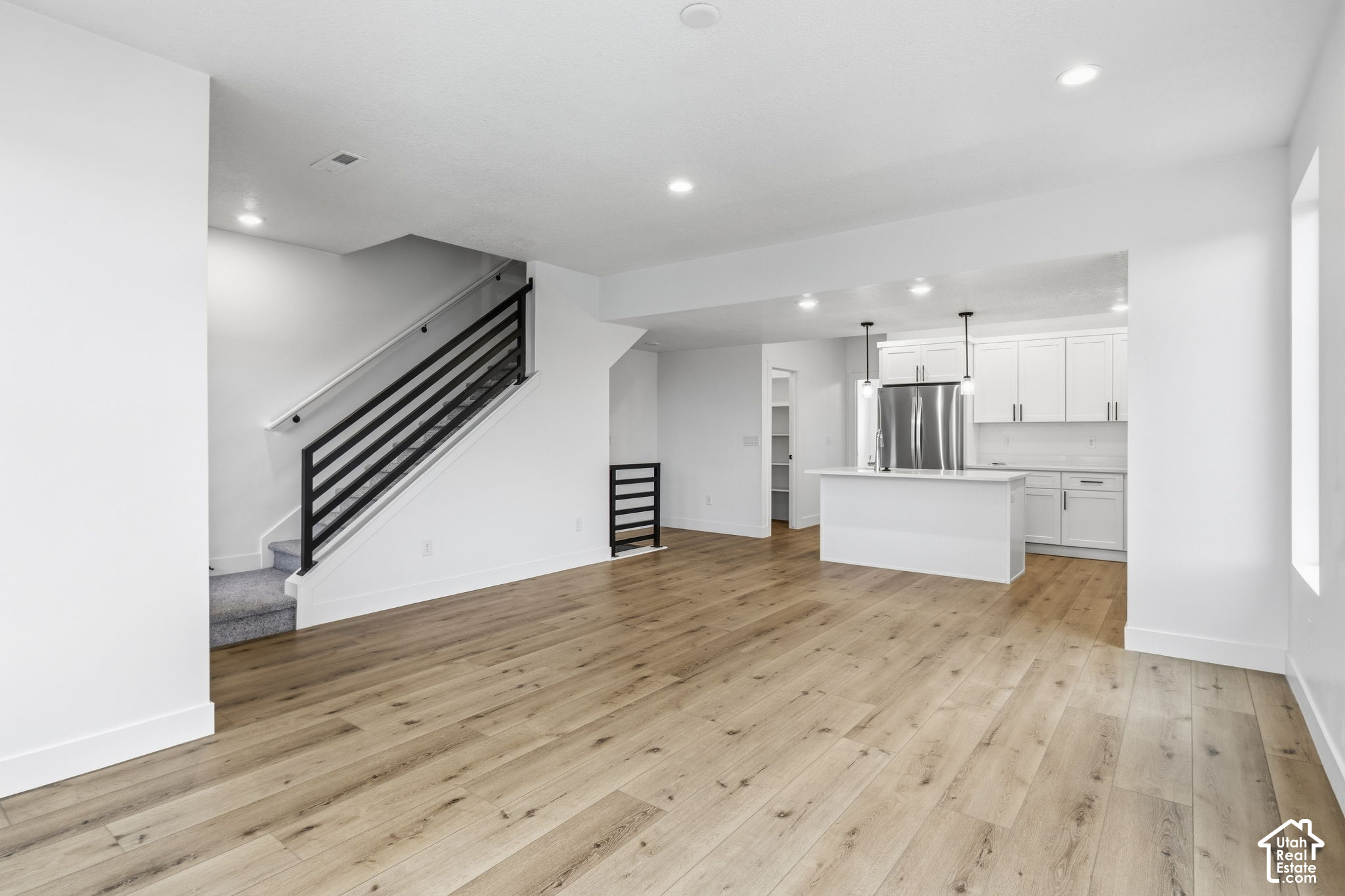 Open living area with wood flooring, white kitchen island, stainless appliances, and a black-railed staircase.