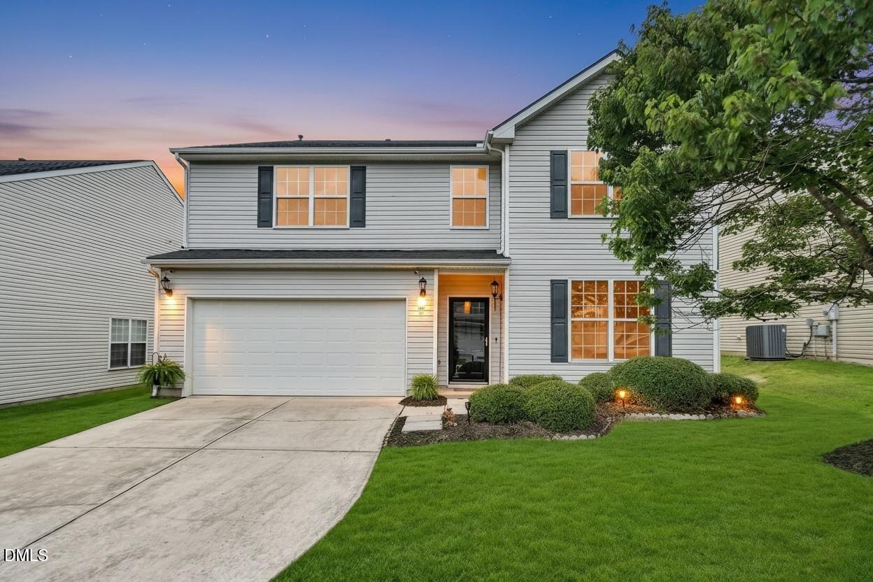 Two-story gray house with a two-car garage, dark shutters, and a landscaped front lawn at dusk.