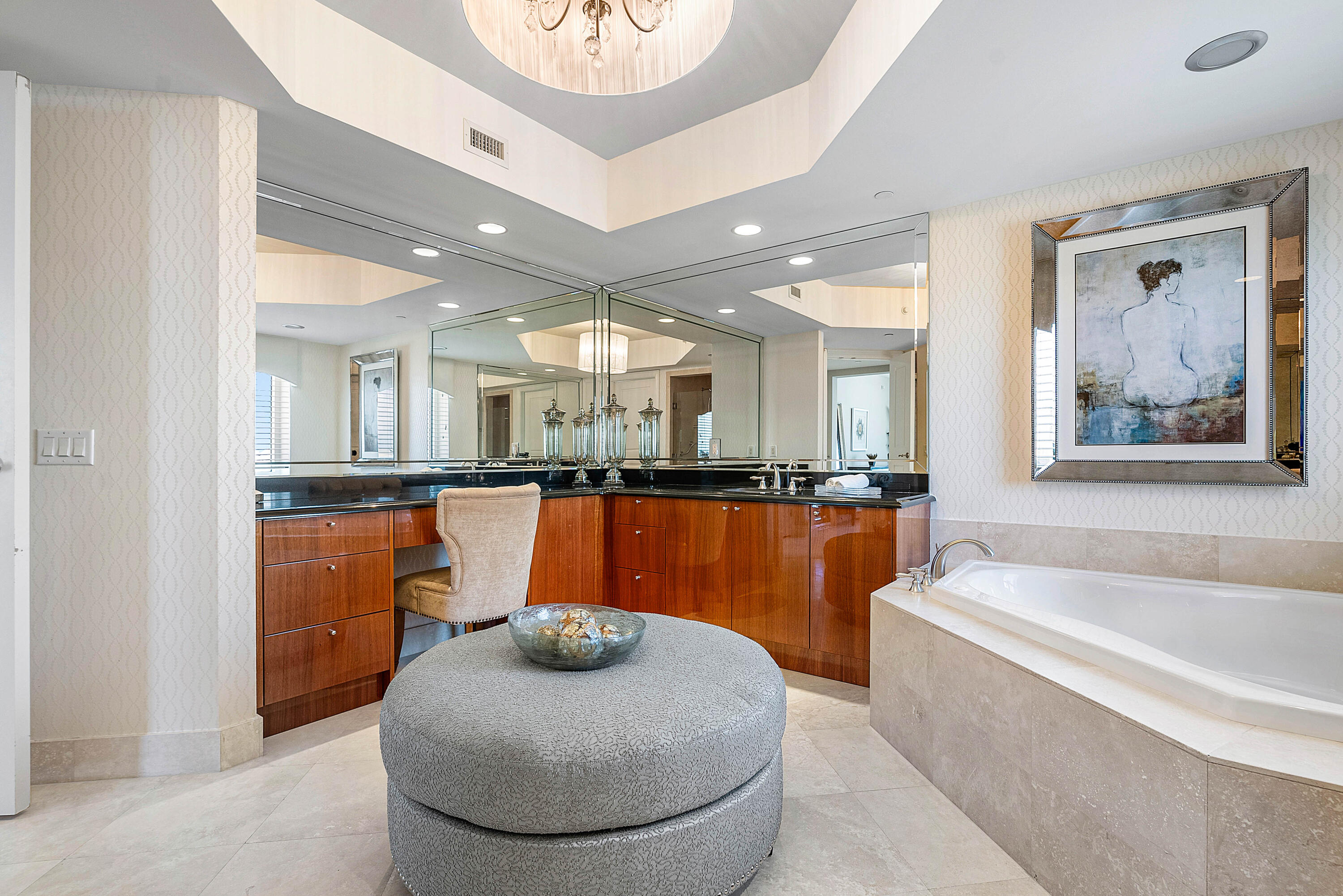 Bathroom with a mirrored vanity, reddish wood cabinetry, built-in soaking tub, and upholstered ottoman.