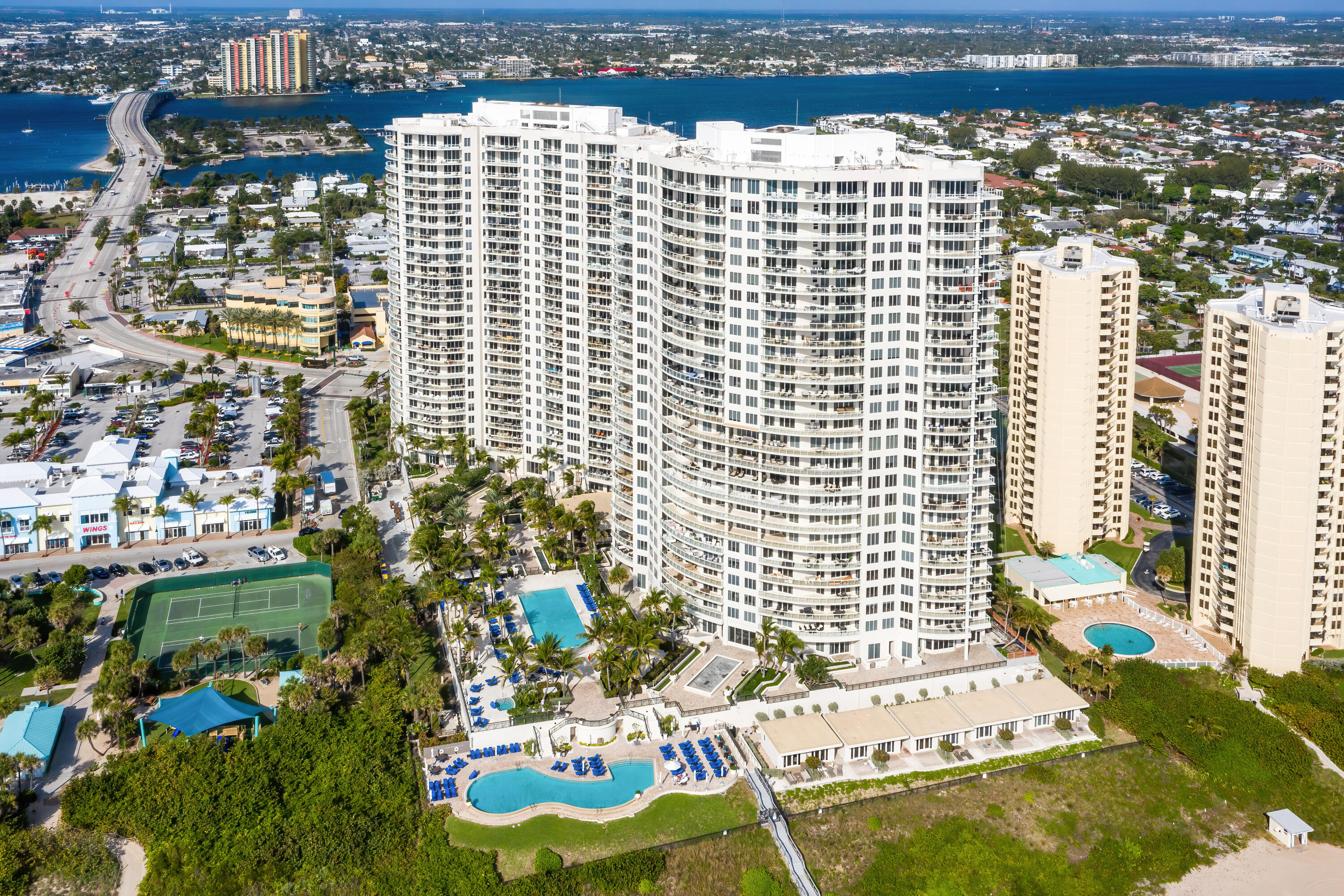 Aerial view of a white high-rise condominium with a pool, tennis court, and waterway beyond.