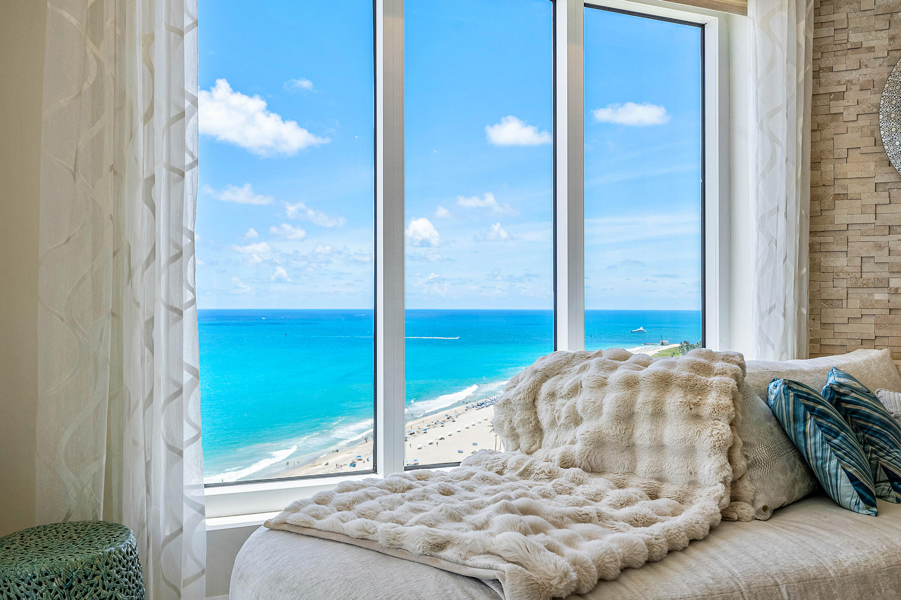 Bedroom daybed with a plush cream throw overlooks turquoise ocean and beach through floor-to-ceiling windows.
