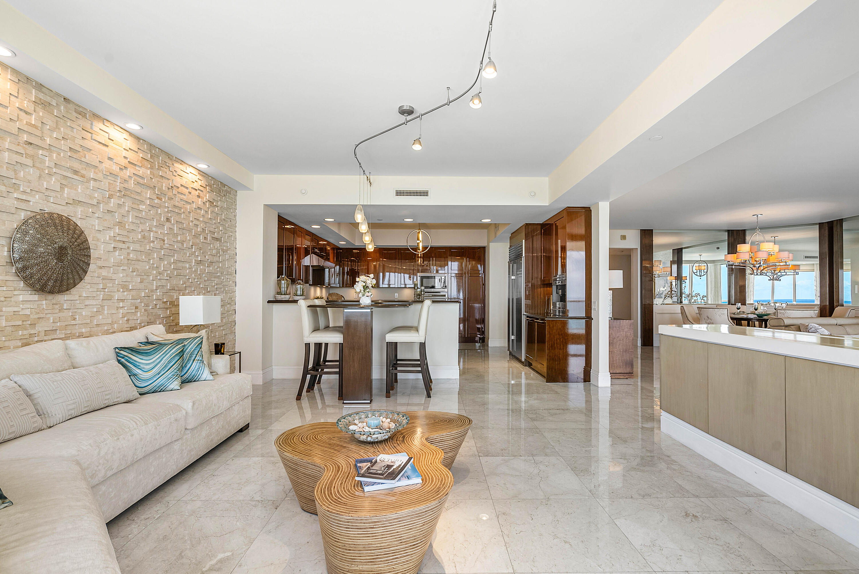 Open-plan condo living room with a stone accent wall, dark wood kitchen cabinetry, and ocean glimpsed through the windows.