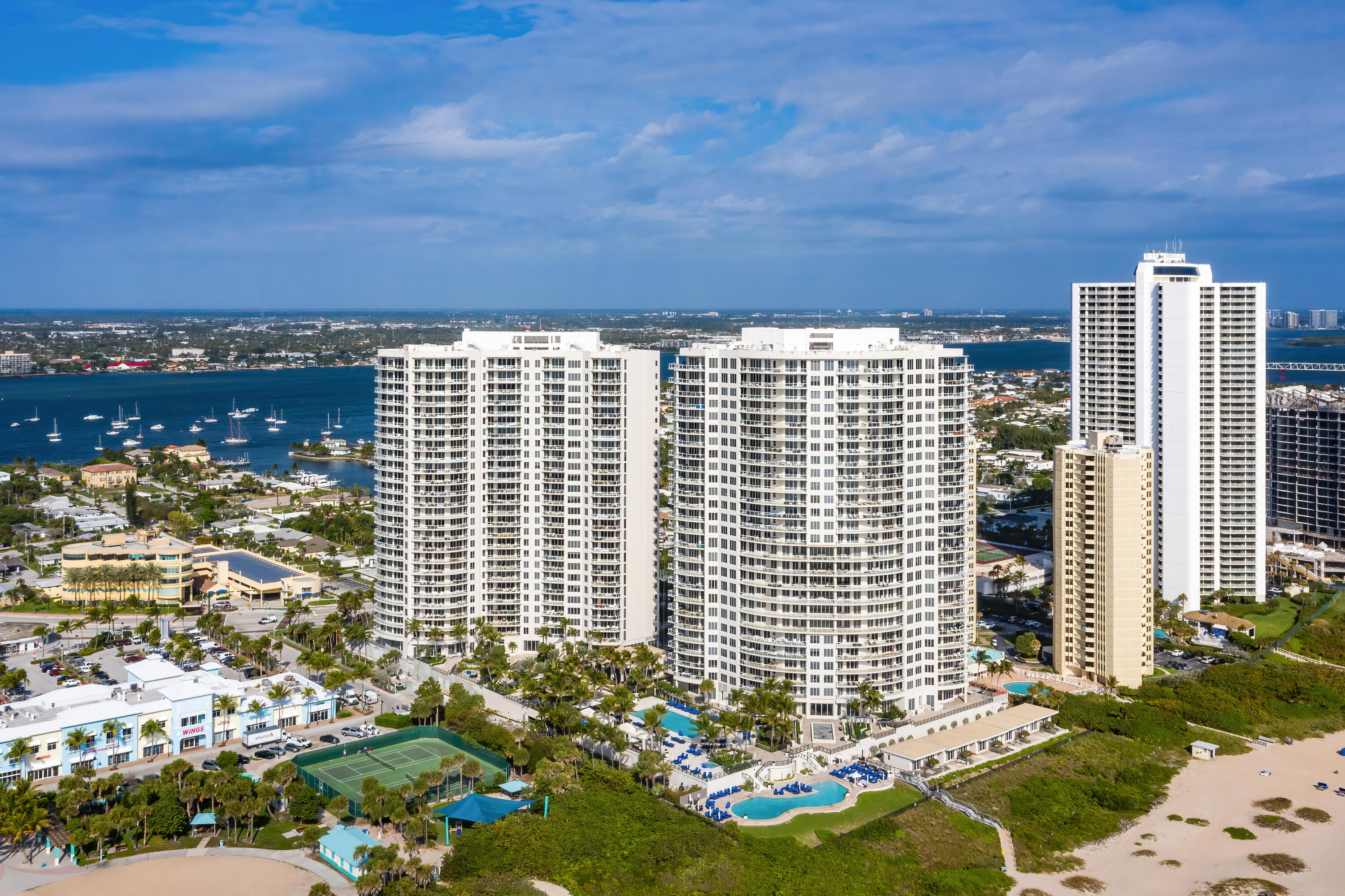 Aerial view of white condominium towers beside a boat-filled waterway, with pools and tennis courts.