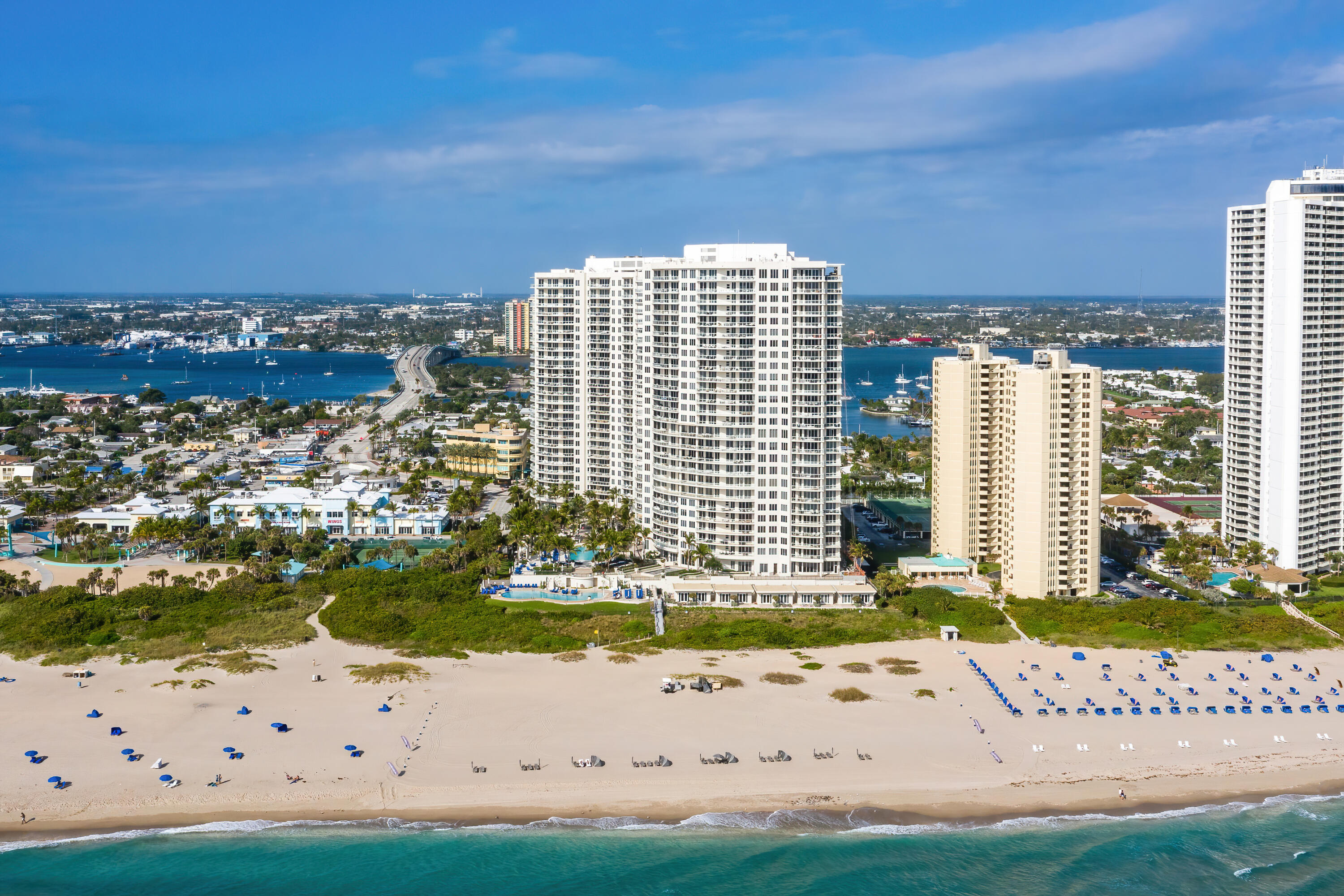 Beachfront condominium towers above a sandy beach, with a curving bridge and waterway in the distance.