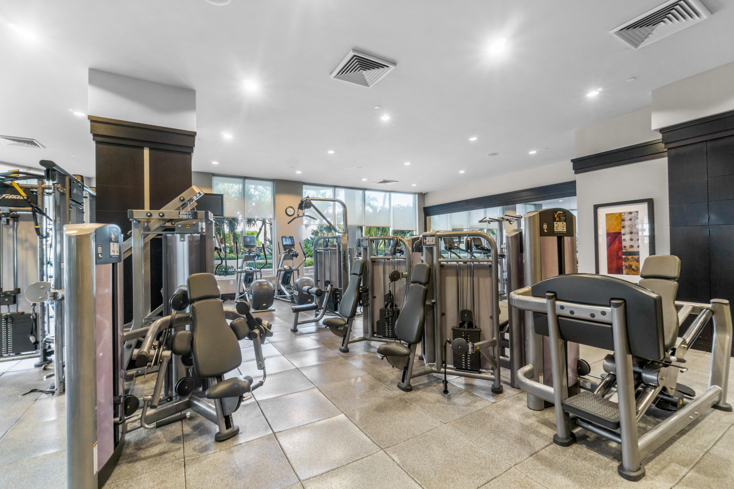 Condominium gym with rows of weight machines, large windows, and a mirrored wall.