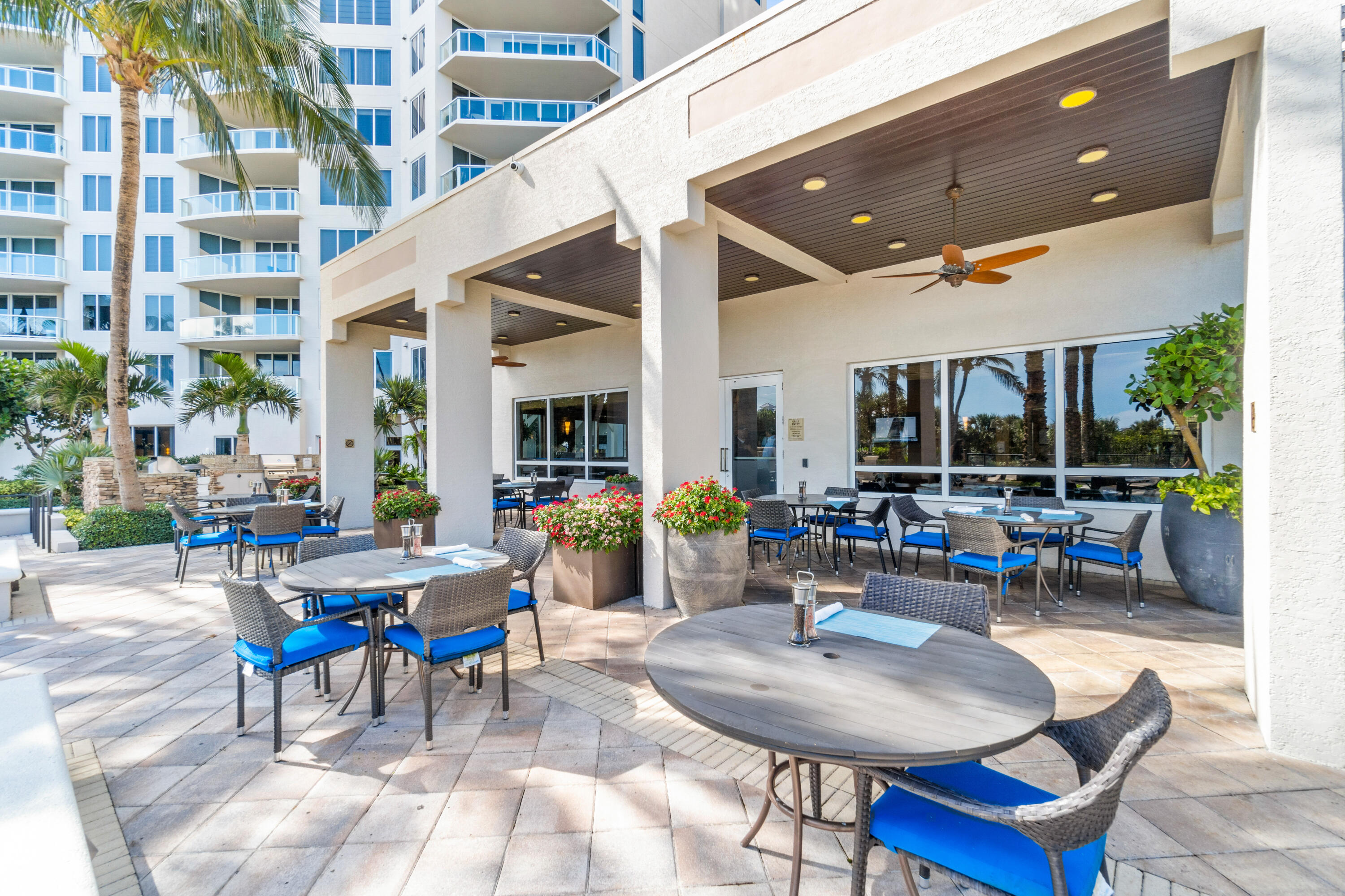 Condominium dining patio with blue-cushioned chairs, ceiling fans, palm trees, and flowering planters.