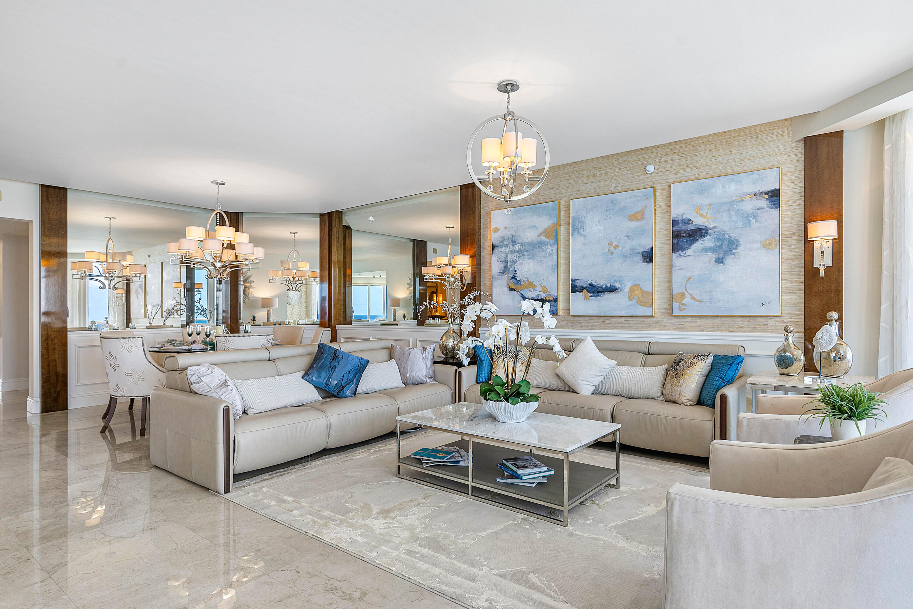 Living room with cream sofas, mirrored walls, blue abstract art, and a marble coffee table.