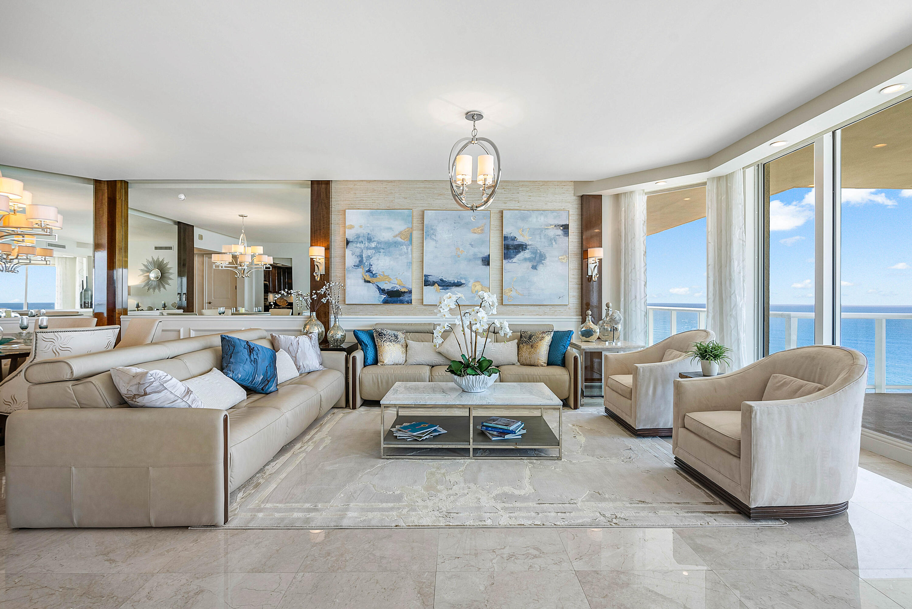 Furnished living room with cream seating, abstract blue artwork, and floor-to-ceiling windows overlooking the ocean.