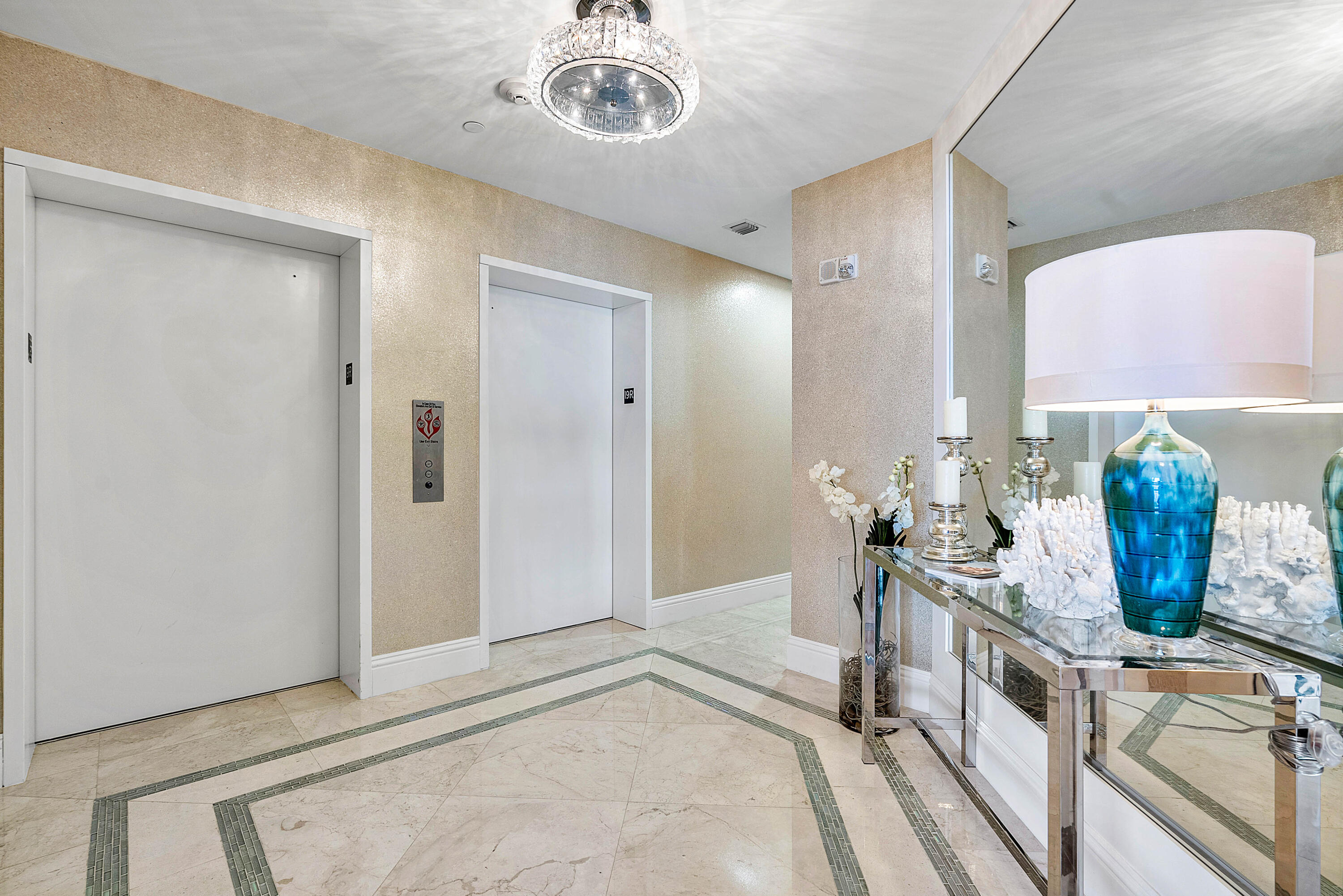 Condominium elevator lobby with two white elevator doors and a mirrored console topped with a blue lamp and flowers.