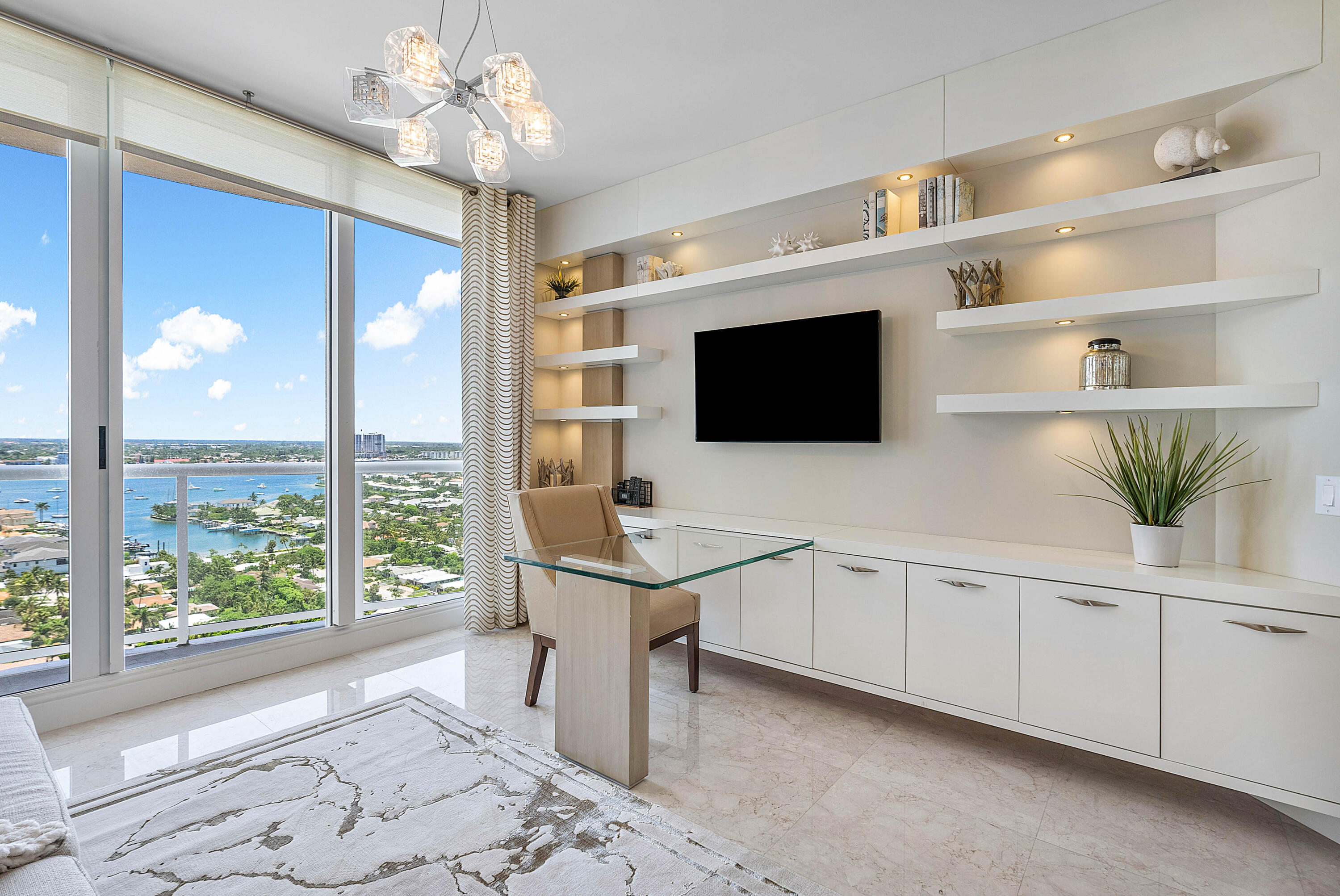 Home office with glass desk and built-in shelves overlooking coastal water and boats.